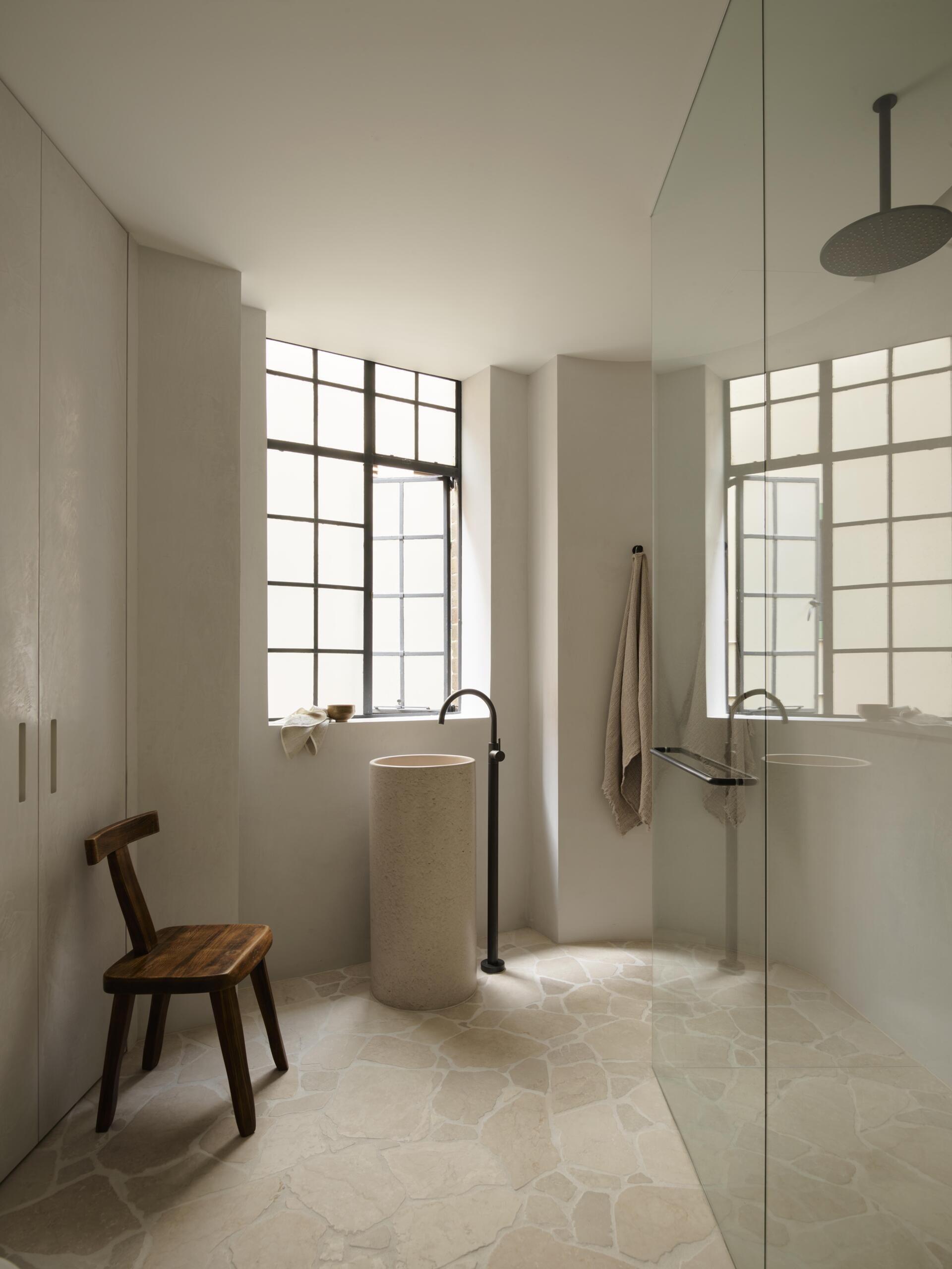 A minimalist bathroom with a cylindrical freestanding stone basin, black floor-mounted faucet, glass shower enclosure, and pale irregular stone floor tiles. Large steel-framed industrial windows bring natural light into the space.