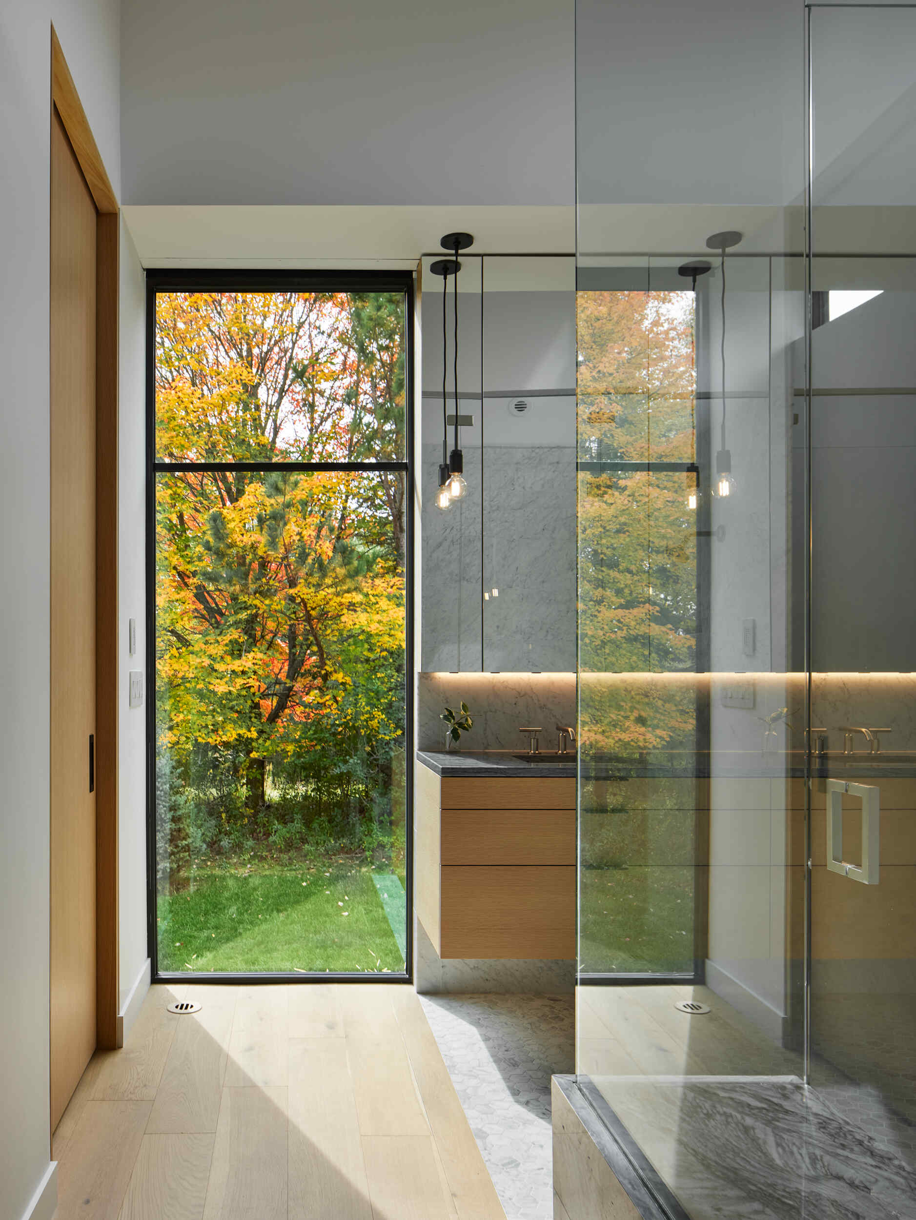 A modern bathroom with a glass-enclosed shower, light wood finishes, and a vanity positioned beside a tall window. The large window provides a view of colorful autumn trees outside.
