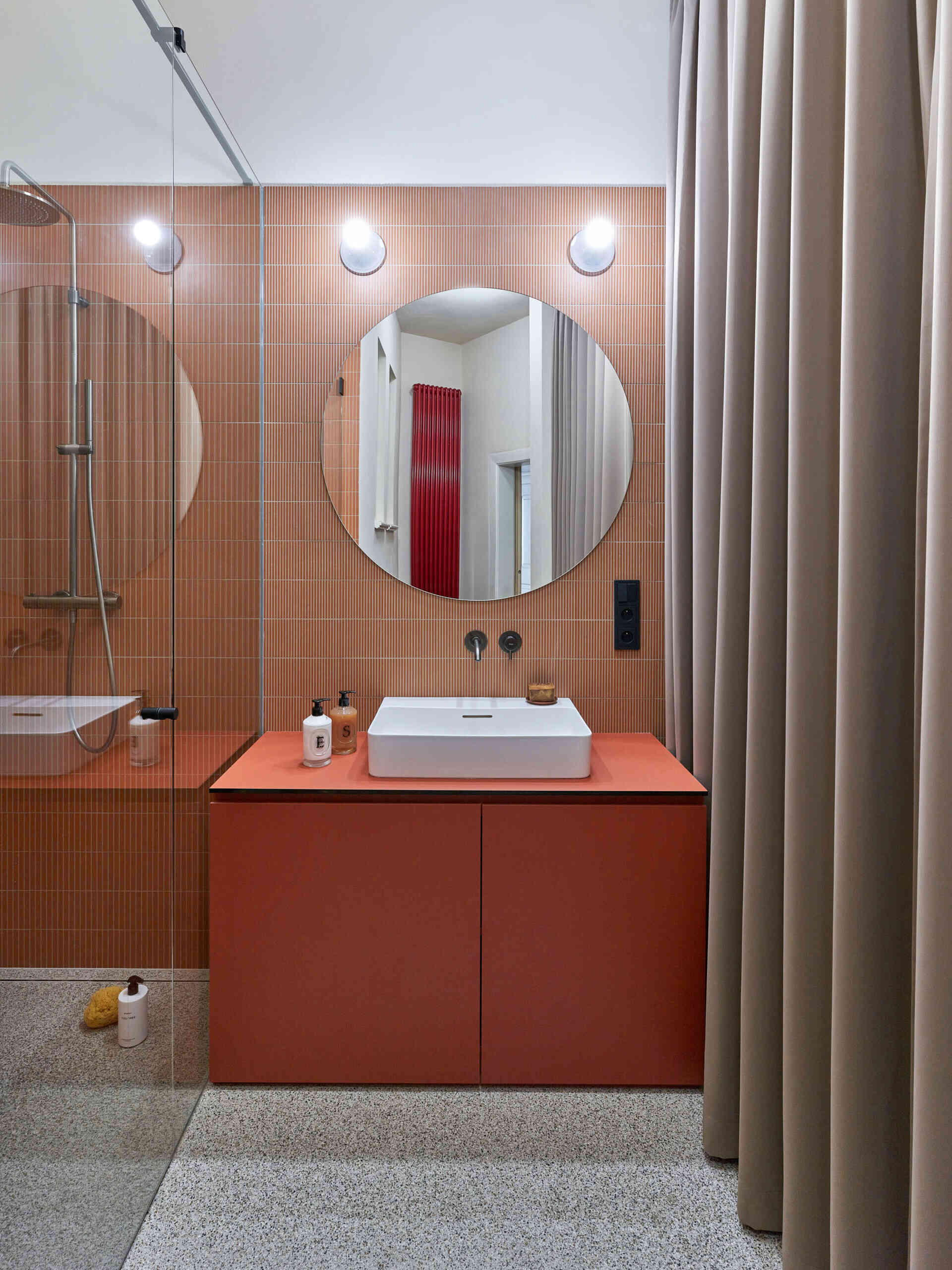 A colorful bathroom with narrow terracotta-colored wall tiles, a matching vanity, white vessel sink, and large round mirror. A glass shower enclosure sits alongside the vanity.