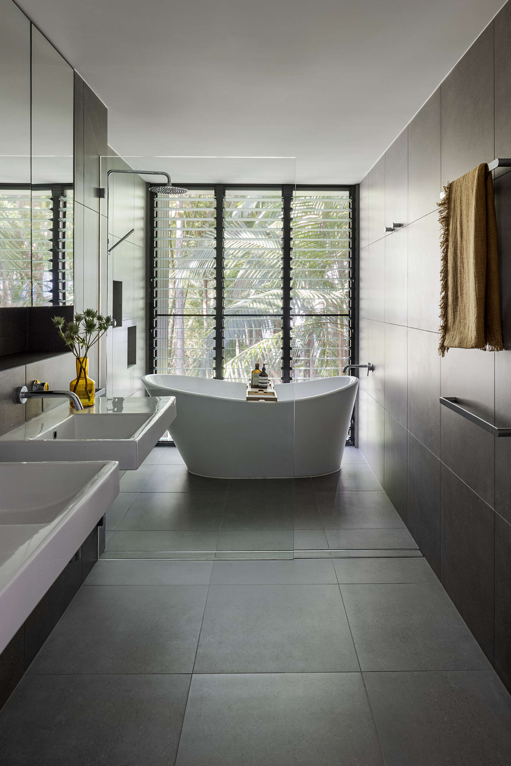 Explore the modern bathrooms at Raintree in Noosa Heads, Australia, designed by Void Building Design. Large-format grey tiles, a freestanding bathtub, minimalist wall-mounted sinks, walk-in showers, and an overhead skylight create bright, contemporary spaces.