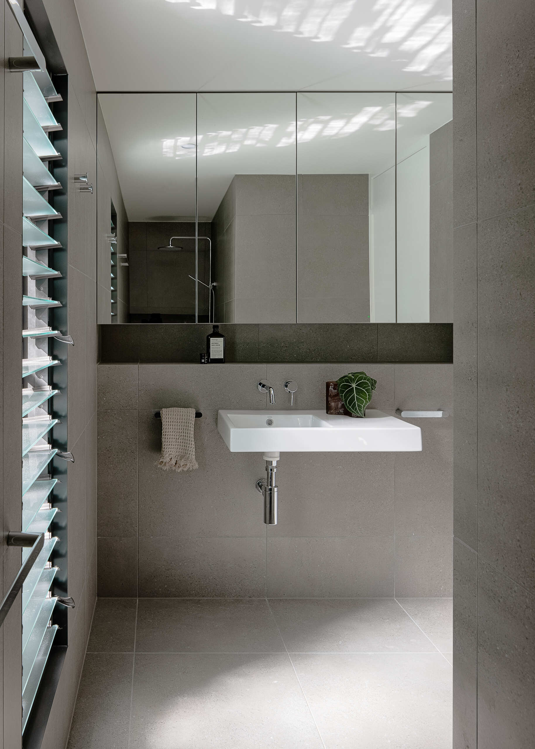 Explore the modern bathrooms at Raintree in Noosa Heads, Australia, designed by Void Building Design. Large-format grey tiles, a freestanding bathtub, minimalist wall-mounted sinks, walk-in showers, and an overhead skylight create bright, contemporary spaces.