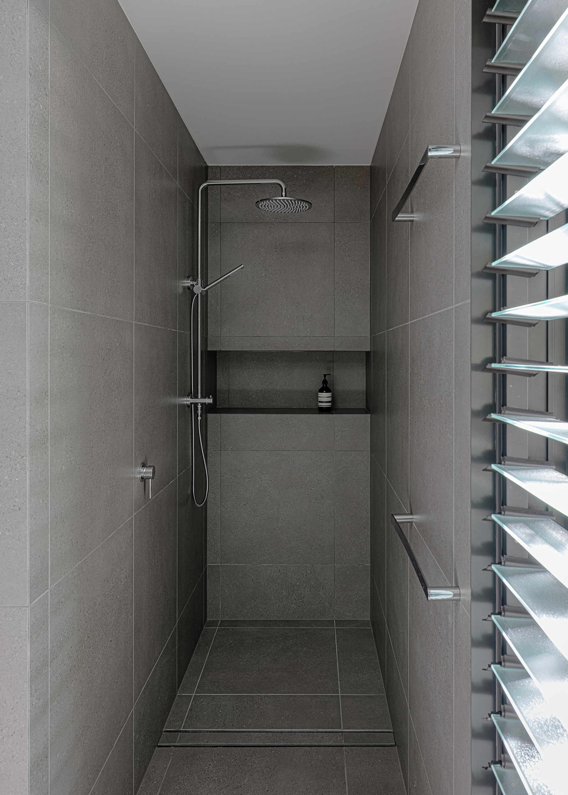 Explore the modern bathrooms at Raintree in Noosa Heads, Australia, designed by Void Building Design. Large-format grey tiles, a freestanding bathtub, minimalist wall-mounted sinks, walk-in showers, and an overhead skylight create bright, contemporary spaces.