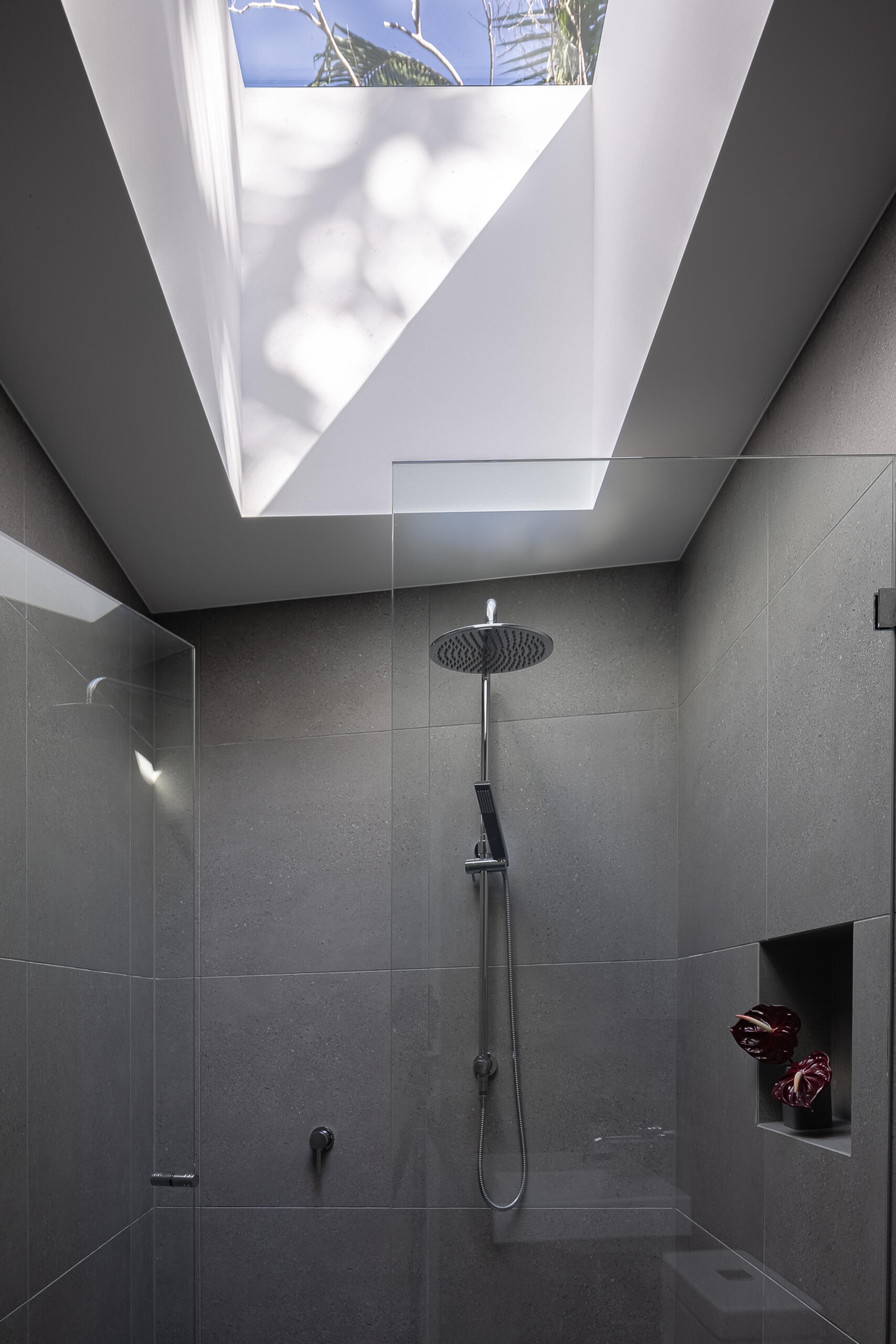 Explore the modern bathrooms at Raintree in Noosa Heads, Australia, designed by Void Building Design. Large-format grey tiles, a freestanding bathtub, minimalist wall-mounted sinks, walk-in showers, and an overhead skylight create bright, contemporary spaces.