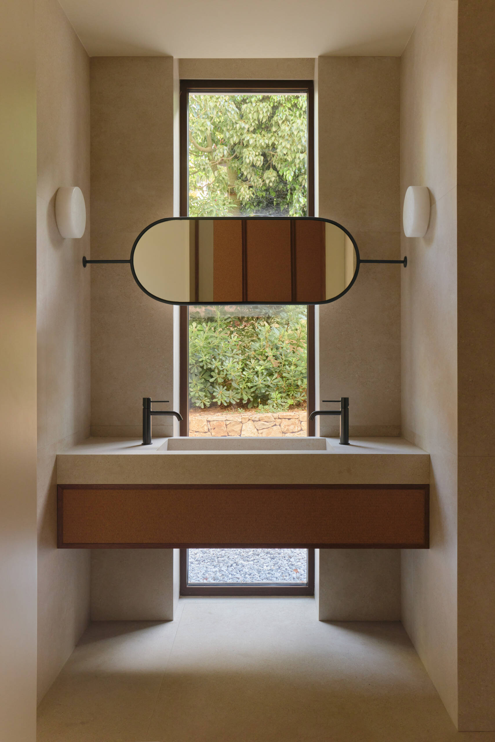 Discover minimalist modern bathrooms with pale stone-look vanities, integrated sinks, oval mirrors, matte black faucets, natural woven cabinetry, and large windows overlooking greenery.
