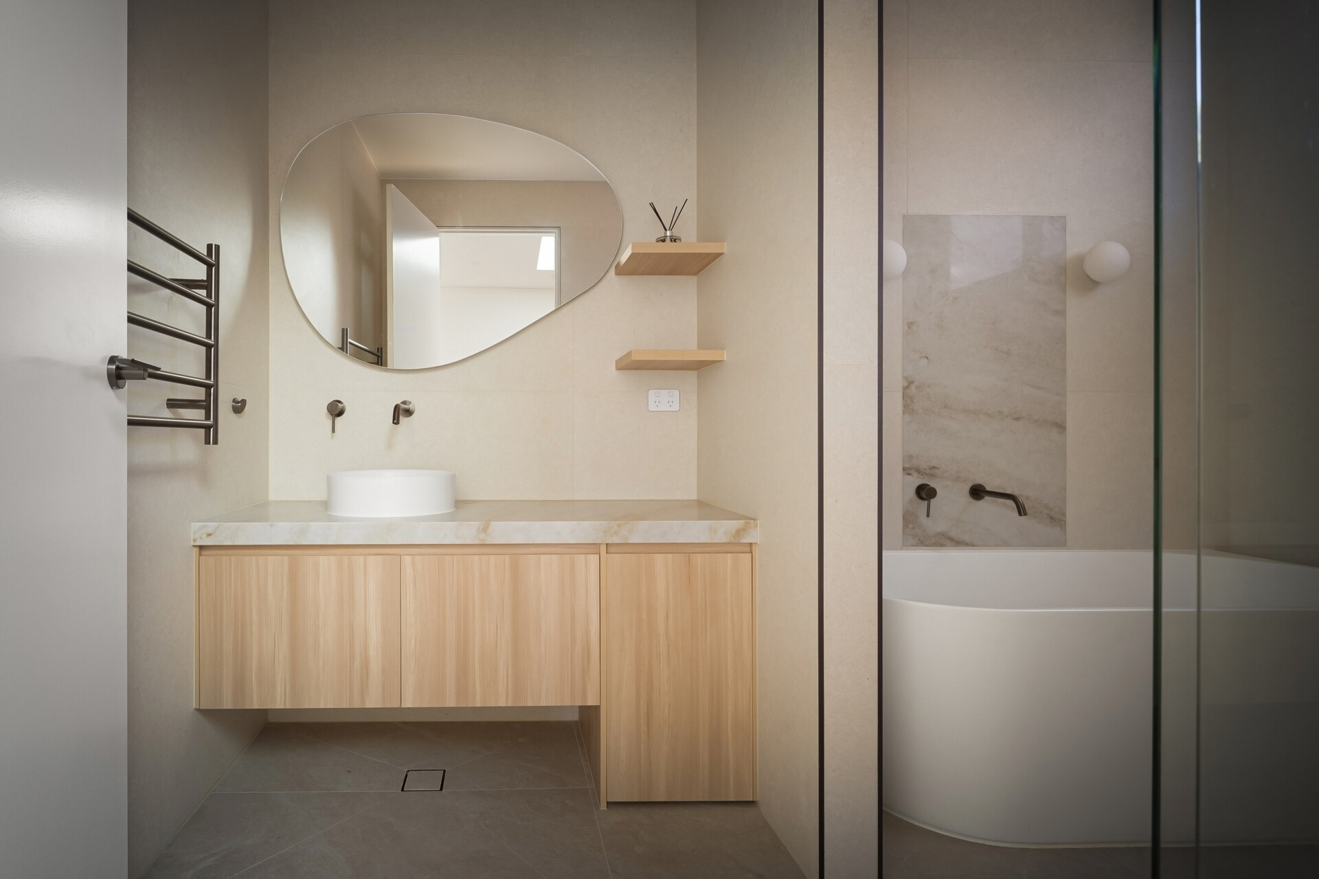 A modern bathroom featuring an irregularly shaped mirror above a floating wood vanity with a white vessel sink and stone countertop. A freestanding bathtub sits beside a stone wall panel with a wall-mounted faucet, while small wood shelves add storage.