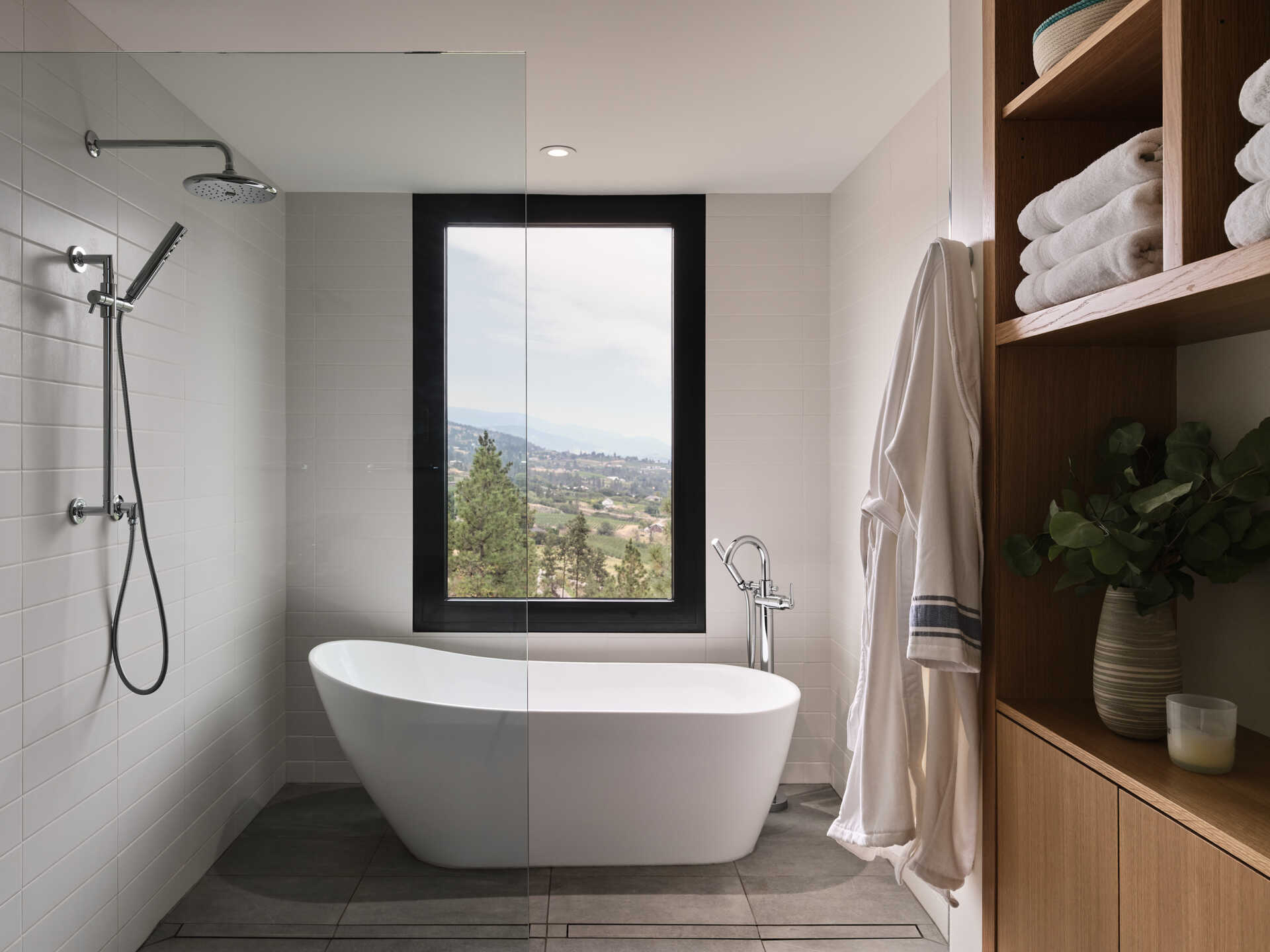 A contemporary bathroom with a white freestanding bathtub positioned beneath a large black-framed window overlooking the hillside. White wall tiles, gray flooring, and open wood shelving complete the space.