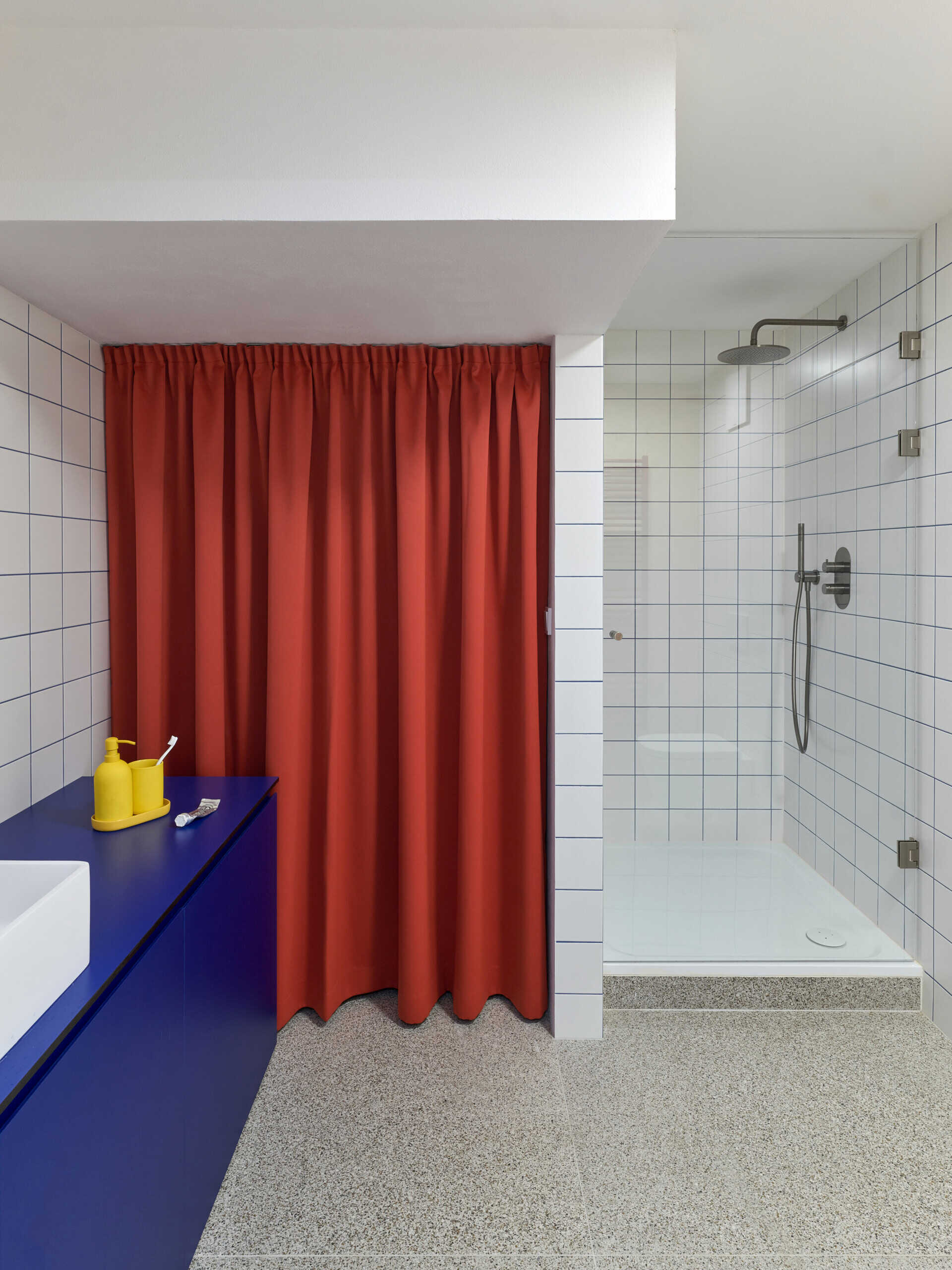 A compact studio bathroom with white square tiles, blue grout lines, a bold blue vanity, and a red curtain. A glass-enclosed shower sits alongside the vanity.