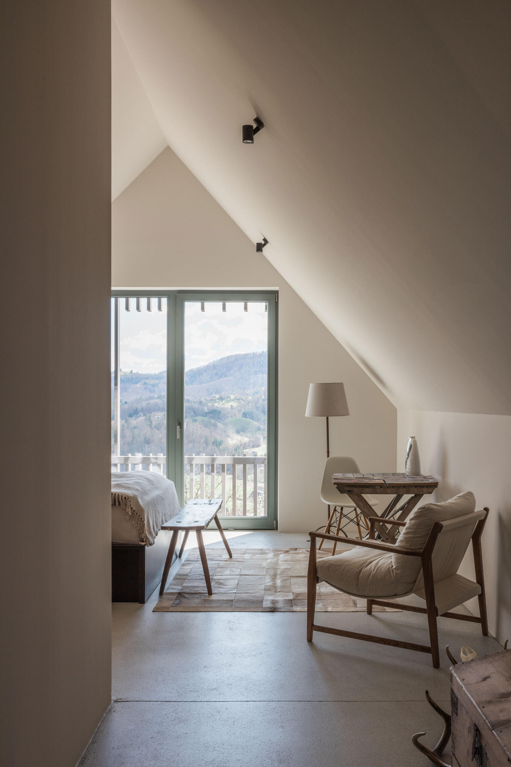 A cosy upper-floor bedroom in a renovated Austrian farmhouse features a sloping ceiling, rustic furniture, a small sitting area, and beautiful countryside views.