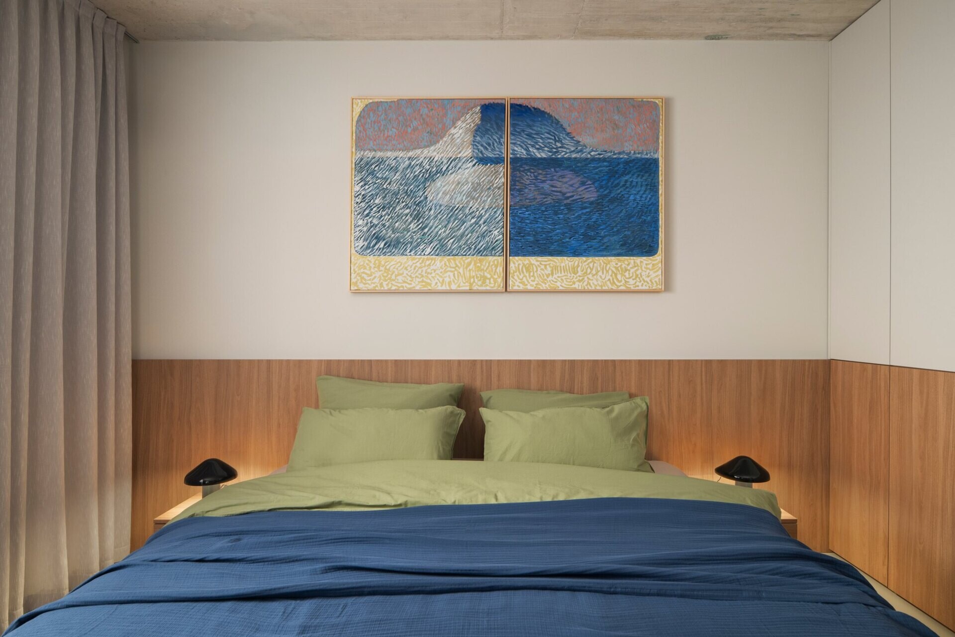 Explore a guest bedroom at Arc House in Istria, Croatia, with a minimalist timber headboard, built-in cabinetry, symmetrical black bedside lamps, blue bedding and colourful two-panel wall art.