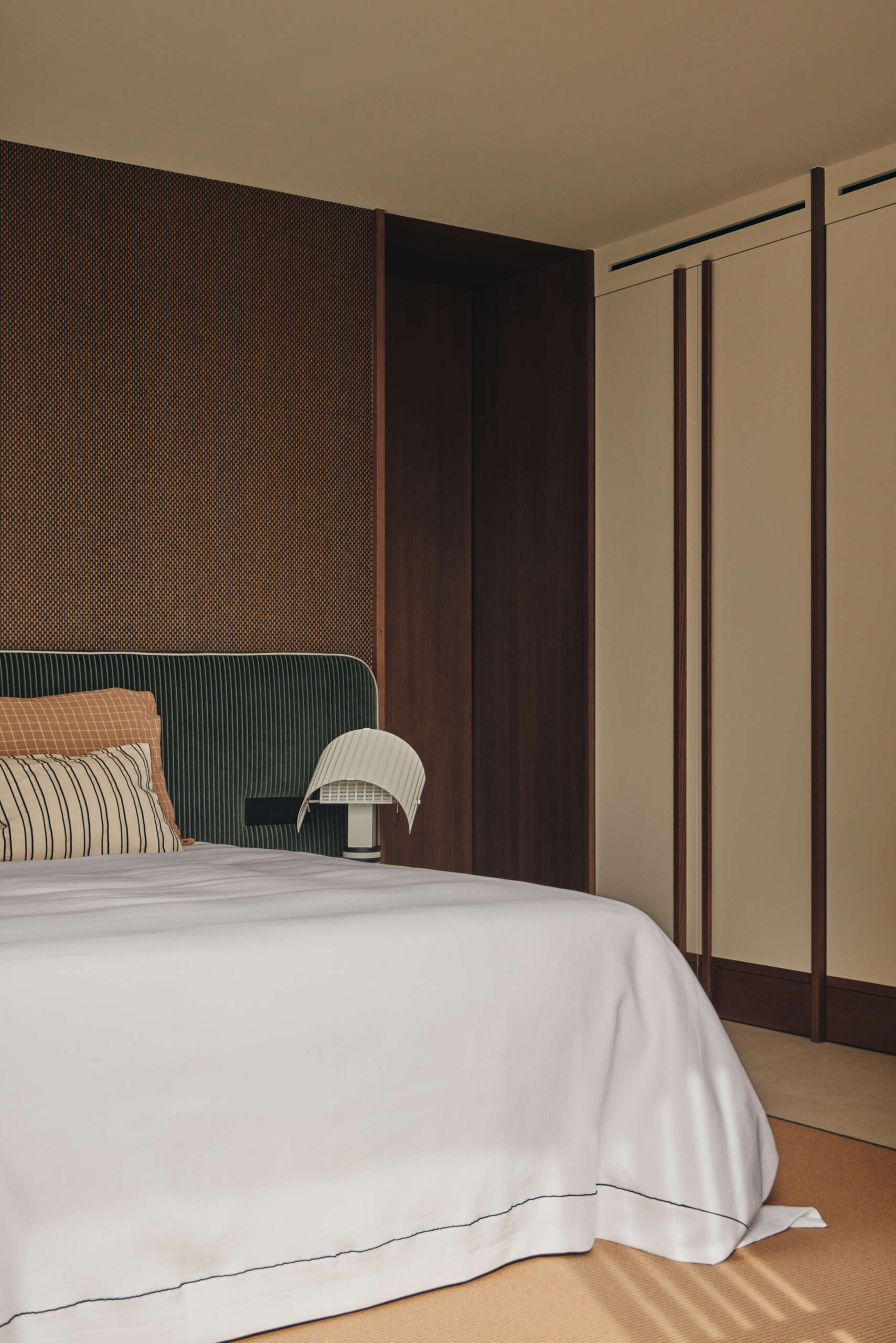 Explore a modern bedroom with a dark green upholstered headboard, sculptural Shogun bedside lamp, natural timber wardrobes, and sliding wooden screens opening onto a flowering garden.