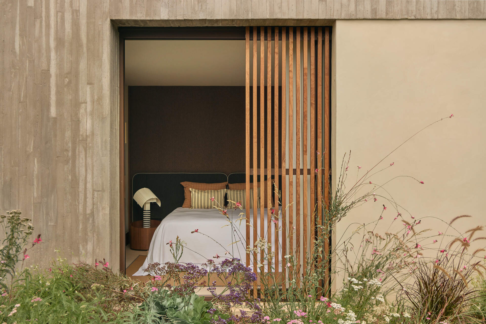 Explore a modern bedroom with a dark green upholstered headboard, sculptural Shogun bedside lamp, natural timber wardrobes, and sliding wooden screens opening onto a flowering garden.
