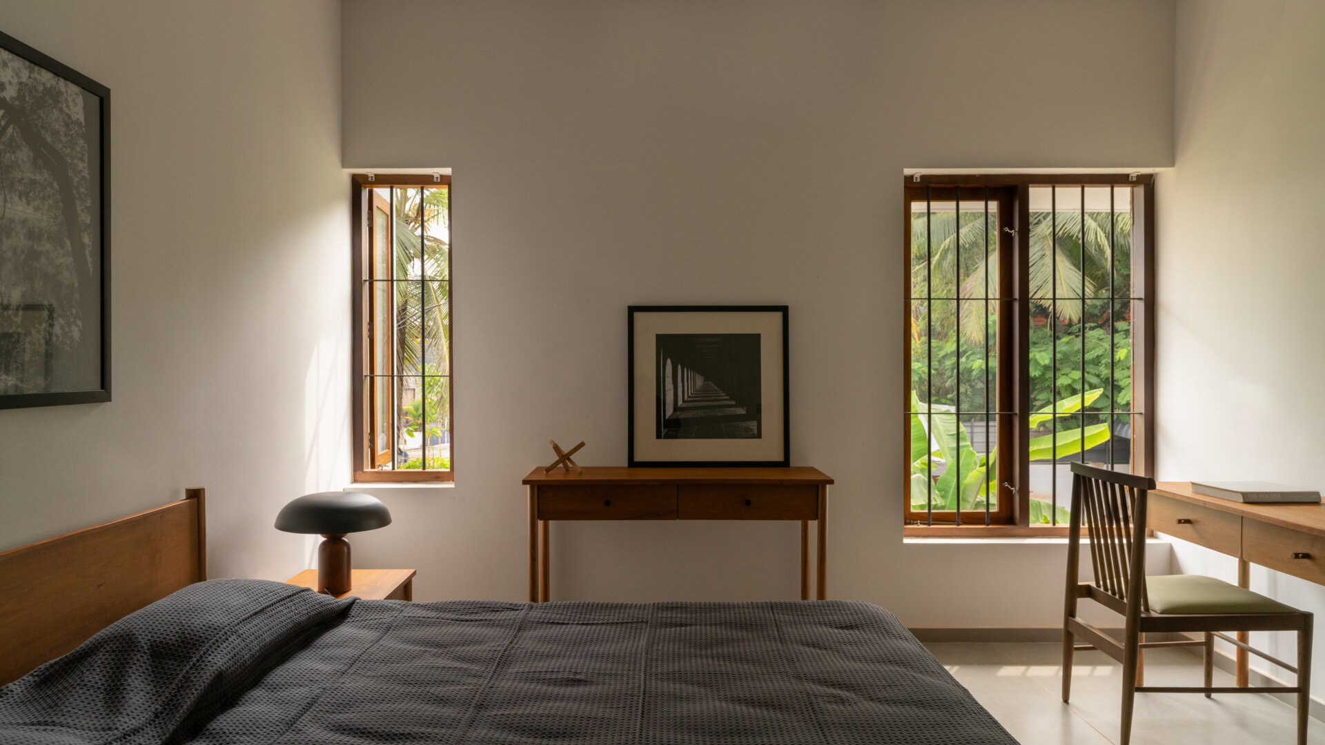 Explore a minimalist bedroom with wooden furniture, charcoal bedding, a small writing desk, and tall windows overlooking tropical greenery in Kerala.
