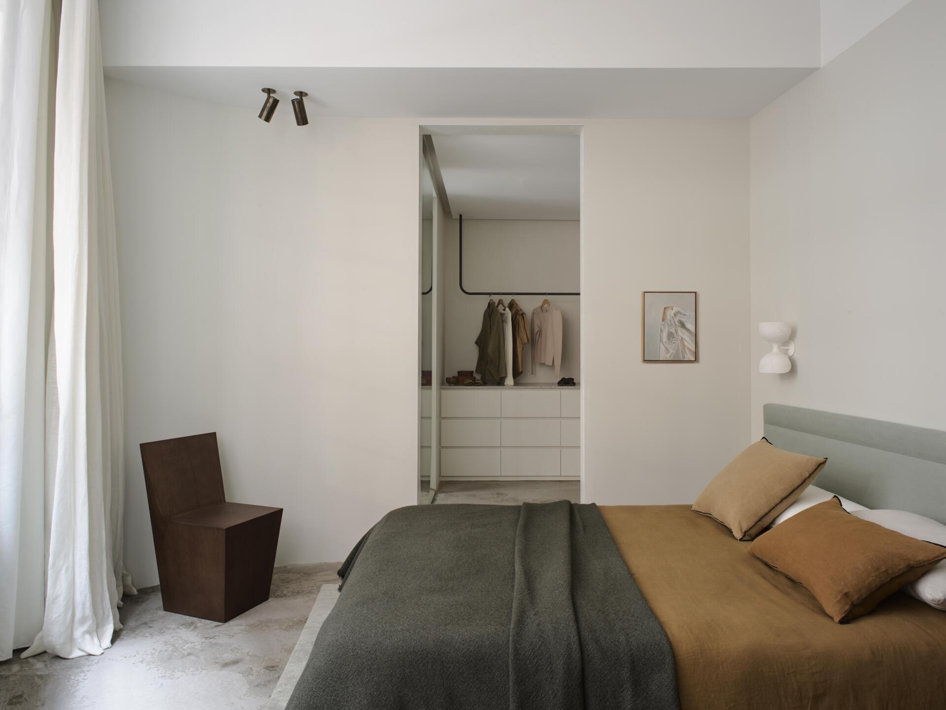 A contemporary minimalist bedroom with a pale upholstered headboard, mustard-colored bedding, dark throw blanket, and sculptural wood chair. White walls, exposed concrete flooring, and an open dressing area create a simple, uncluttered interior.
