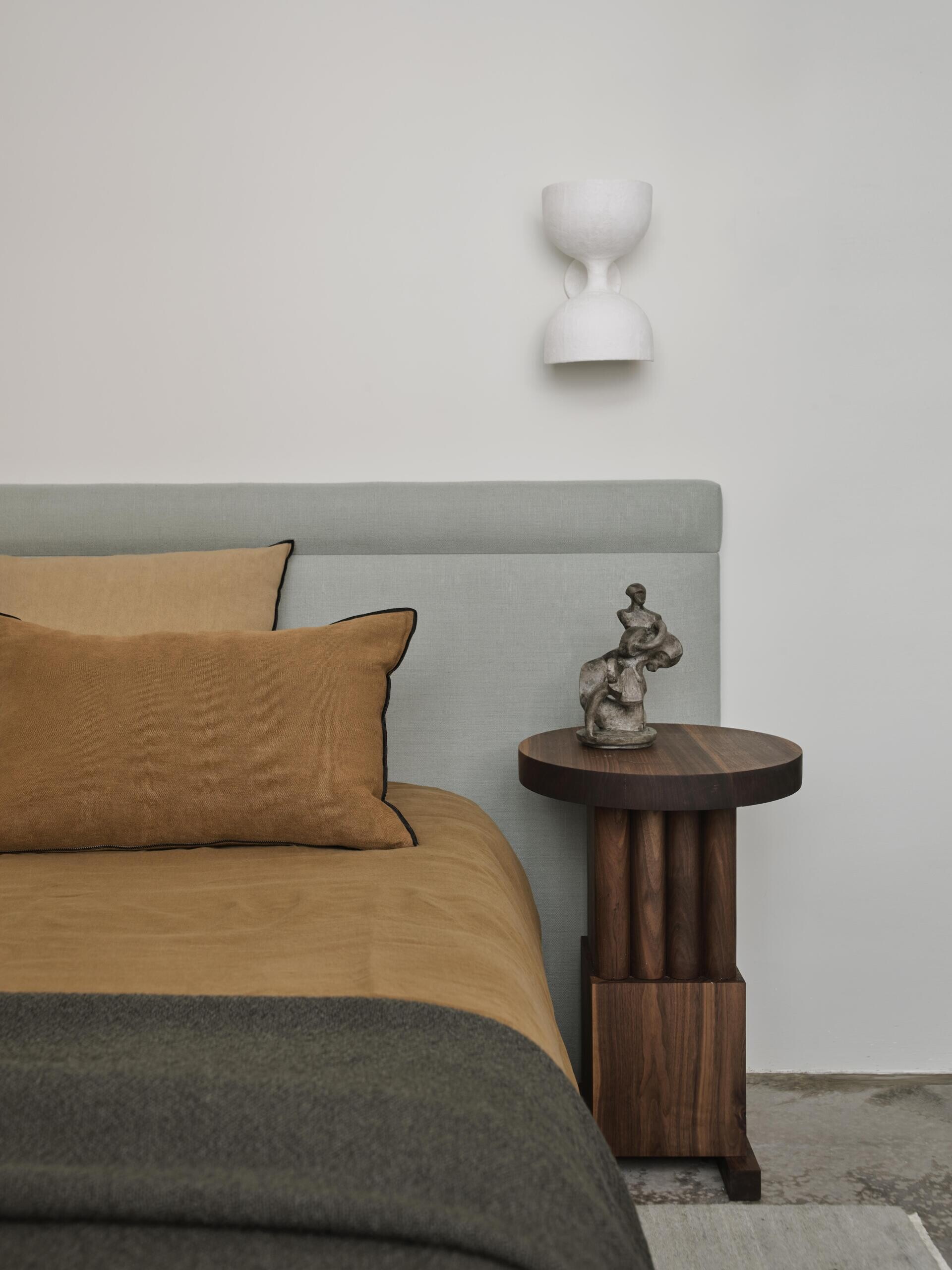 A contemporary minimalist bedroom with a pale upholstered headboard, mustard-colored bedding, dark throw blanket, and sculptural wood chair. White walls, exposed concrete flooring, and an open dressing area create a simple, uncluttered interior.