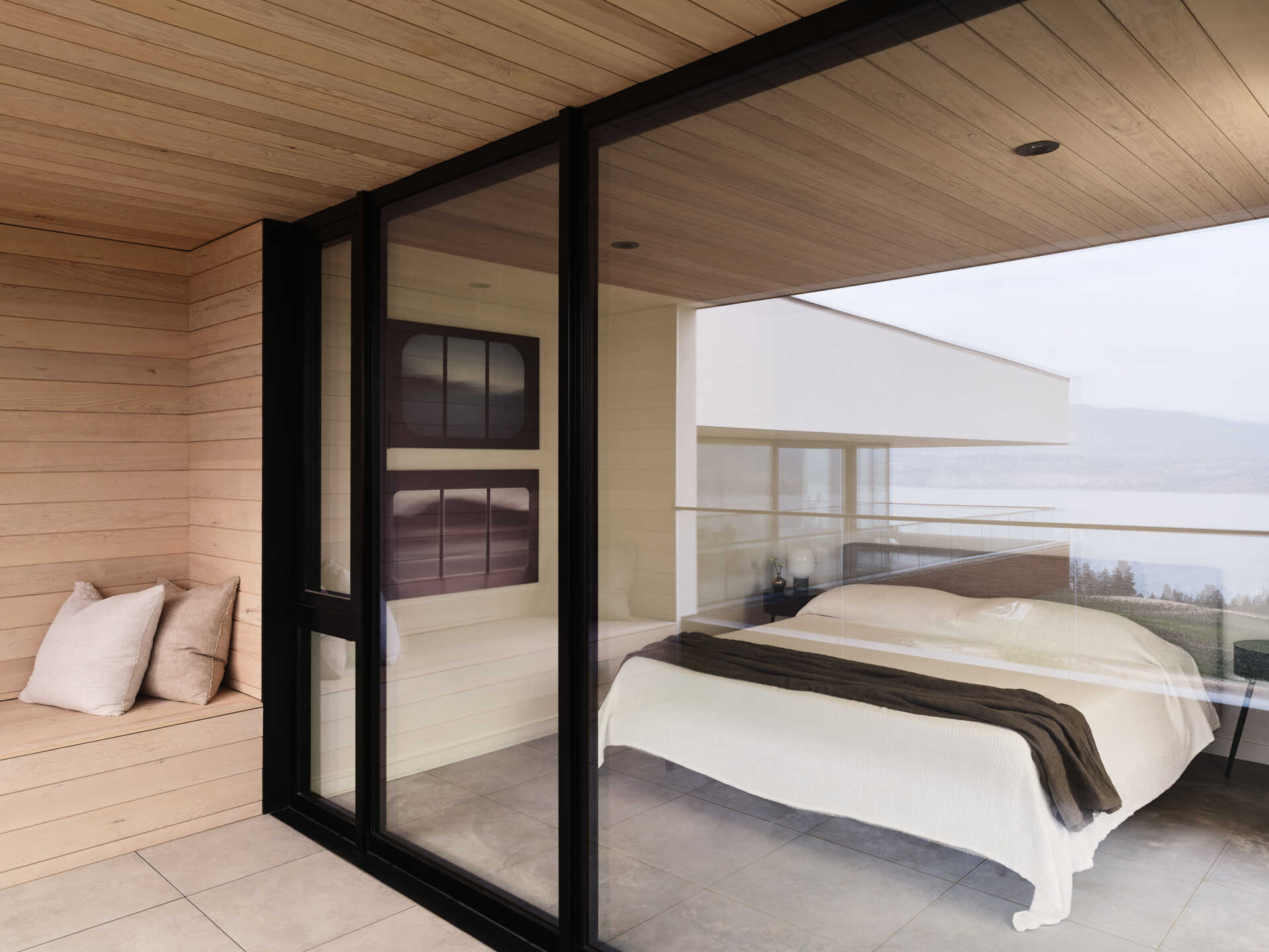 A modern bedroom opens through black-framed glass doors onto a covered terrace with built-in wood seating. Warm wood ceilings, neutral bedding, and large windows create a simple interior overlooking the landscape.