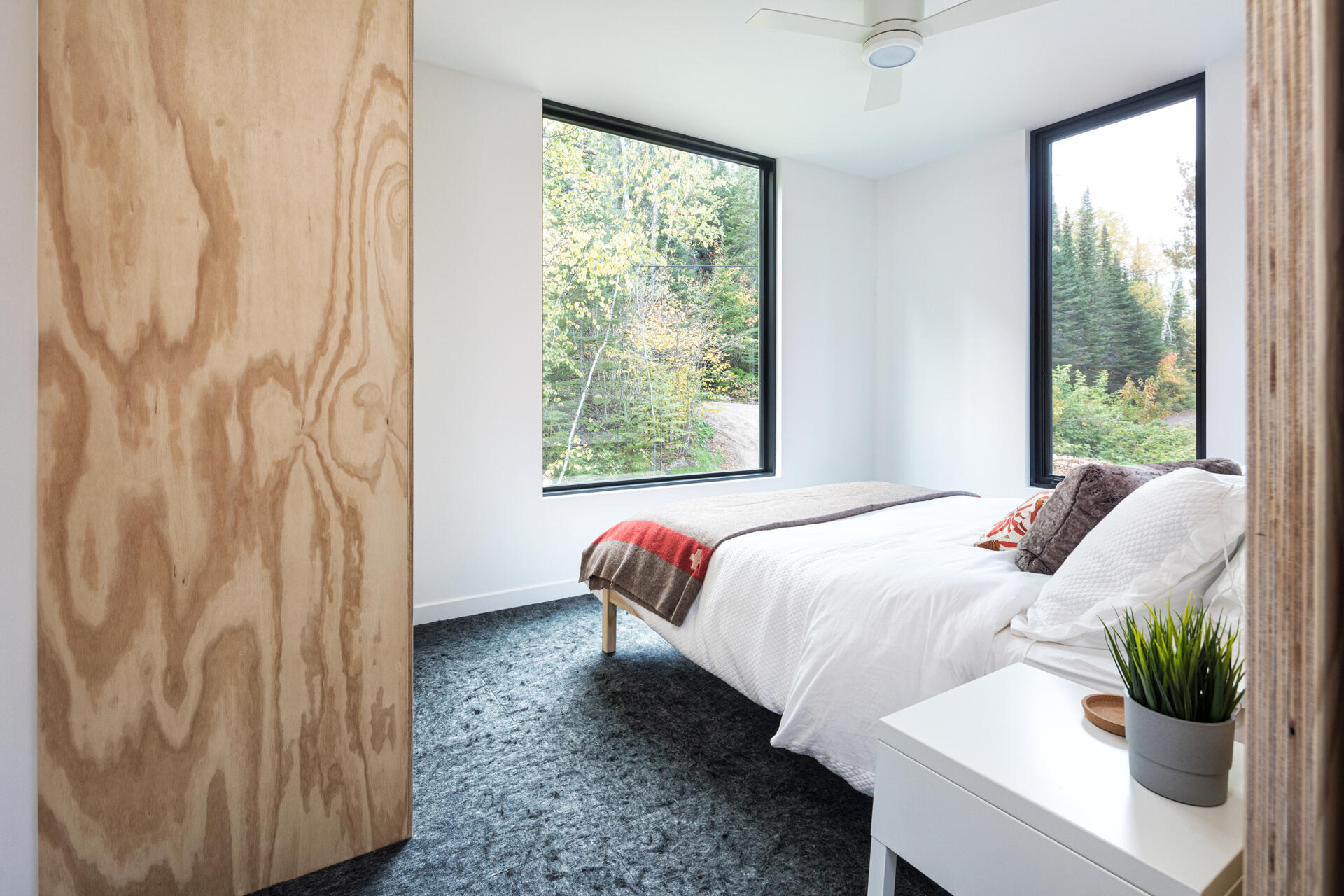 A minimalist cabin bedroom with white walls, a plywood closet, dark gray carpet, and large black-framed windows overlooking the forest. A simple bed with white bedding occupies the compact space.