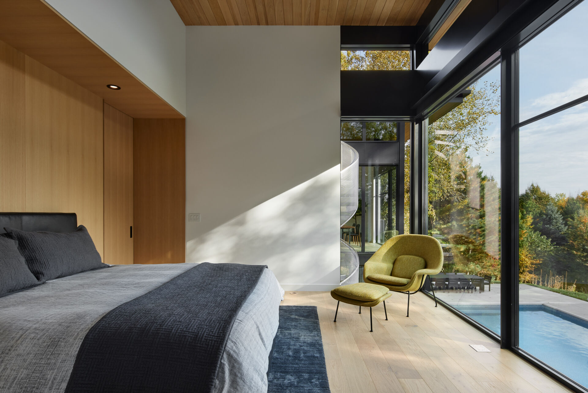 A modern primary bedroom with a gray upholstered bed, built-in wood cabinetry, and a green lounge chair beside floor-to-ceiling windows. The glass overlooks the swimming pool and surrounding trees.