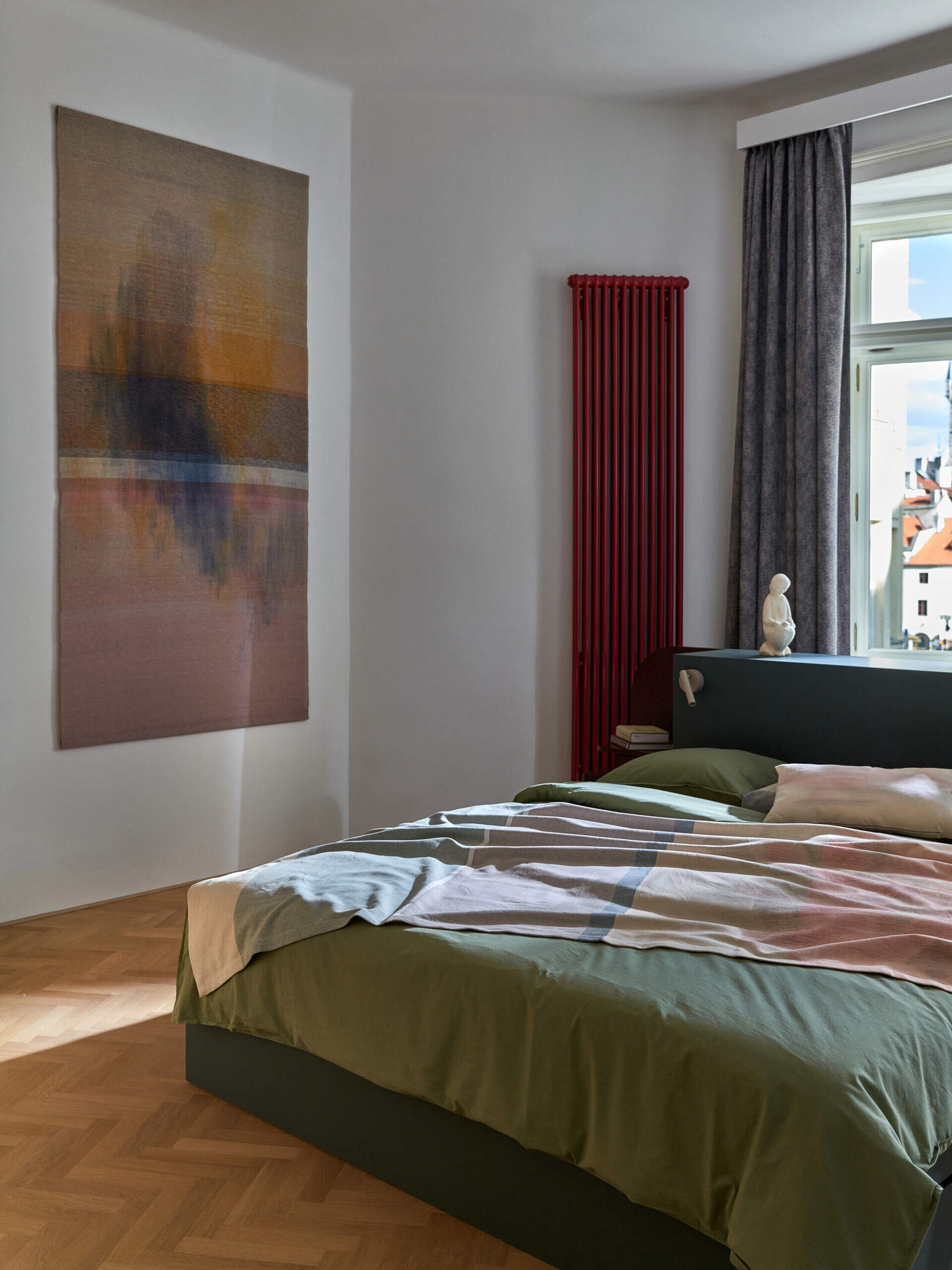 A contemporary bedroom with a stone-green bed frame, green bedding, a red radiator, and a large abstract wall hanging. A traditional window frames a view over Prague's rooftops.