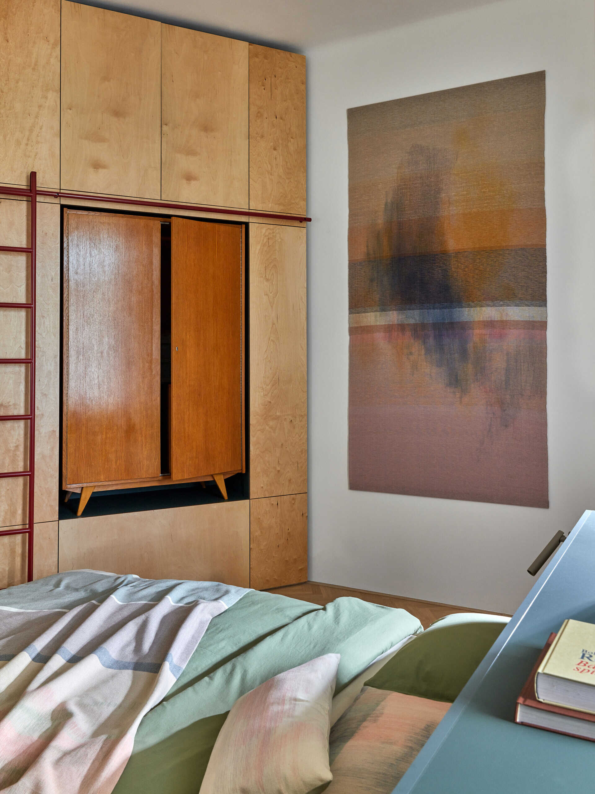 A modern bedroom combines a stone-green bed with pale built-in cabinets and a warm-toned vintage-style cabinet integrated into the storage wall. A red ladder and abstract textile artwork add color.