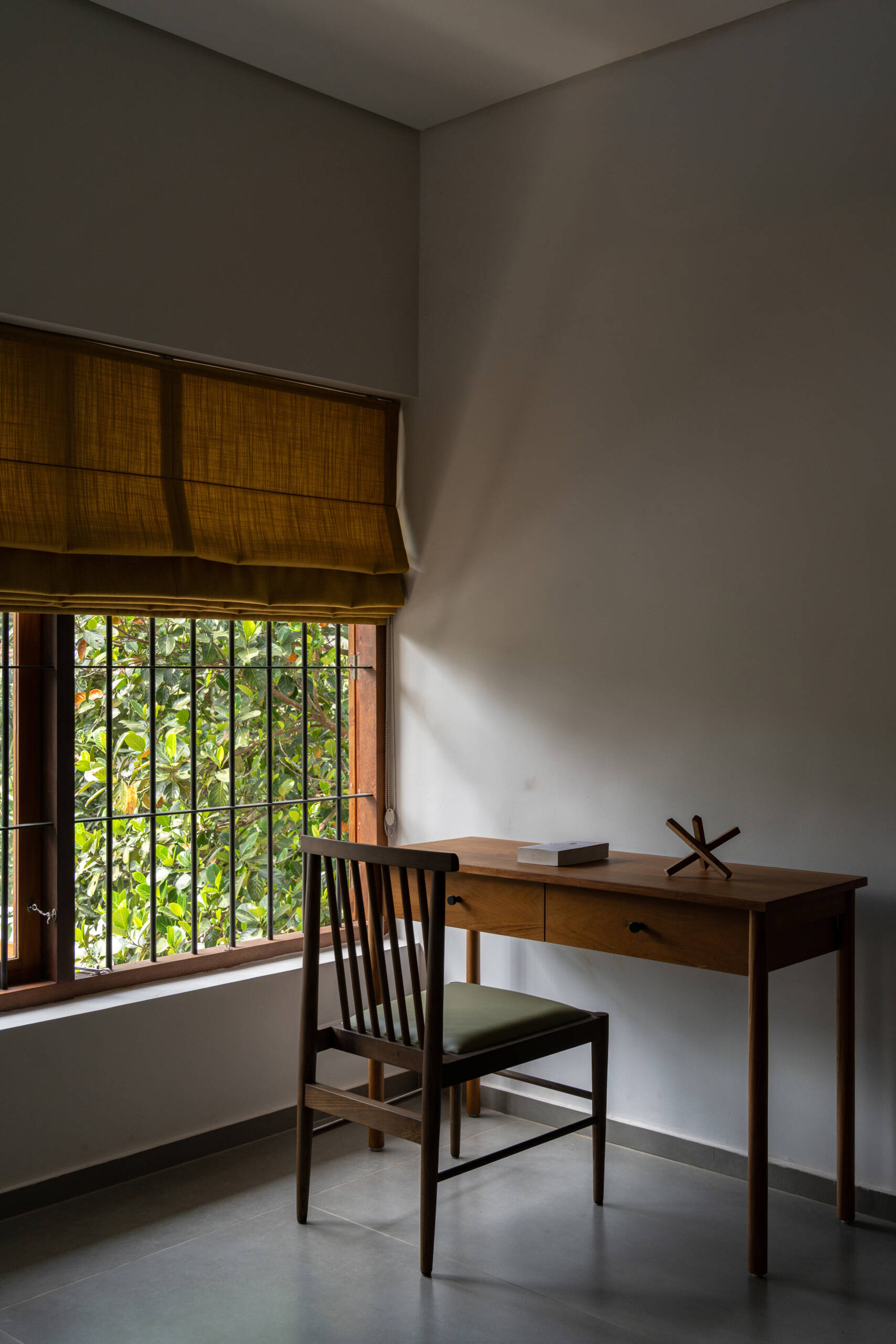 Explore a minimalist bedroom with wooden furniture, charcoal bedding, a small writing desk, and tall windows overlooking tropical greenery in Kerala.
