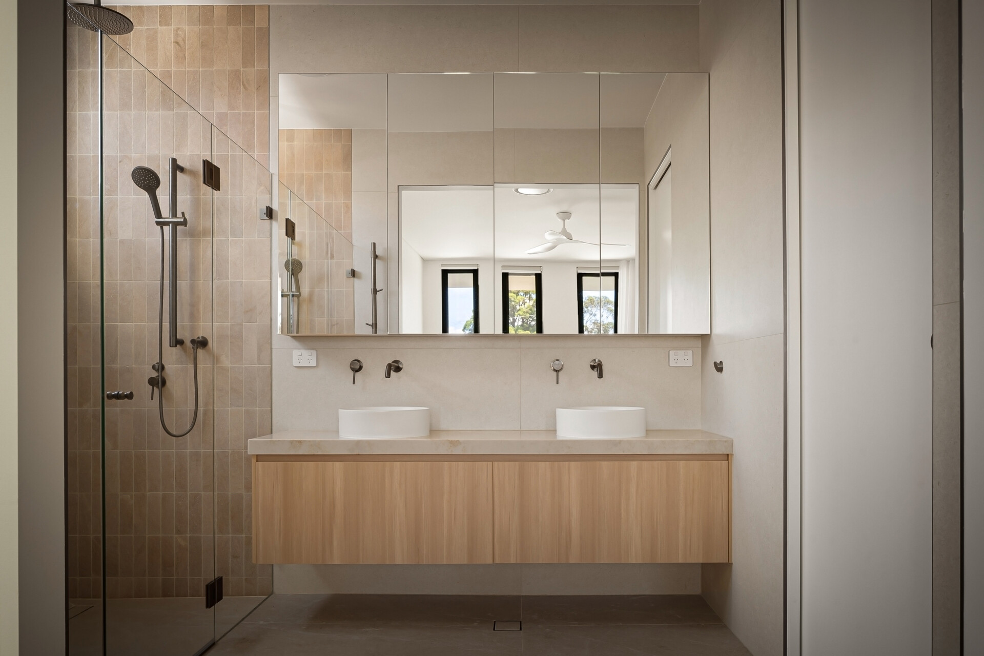 A contemporary ensuite bathroom with a floating double wood vanity, two white vessel sinks, and a wide mirrored cabinet. The walk-in shower features narrow beige wall tiles, frameless glass panels, and dark metal fixtures.