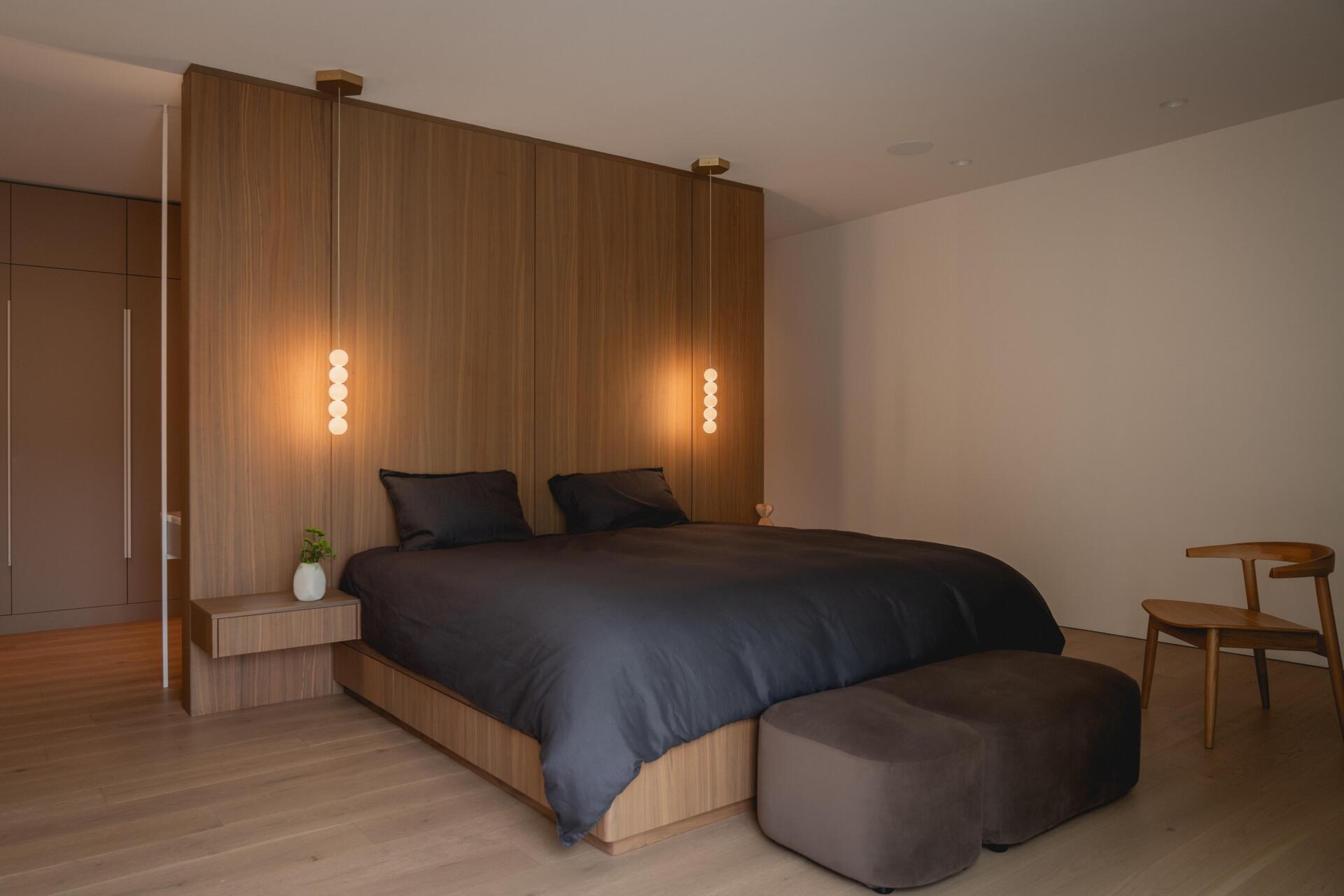 Discover a minimalist bedroom inside The Lens, a modern Montreal home by MU Architecture. A full-height walnut headboard, floating bedside tables, delicate pendant lights, and charcoal bedding create a warm contemporary interior.