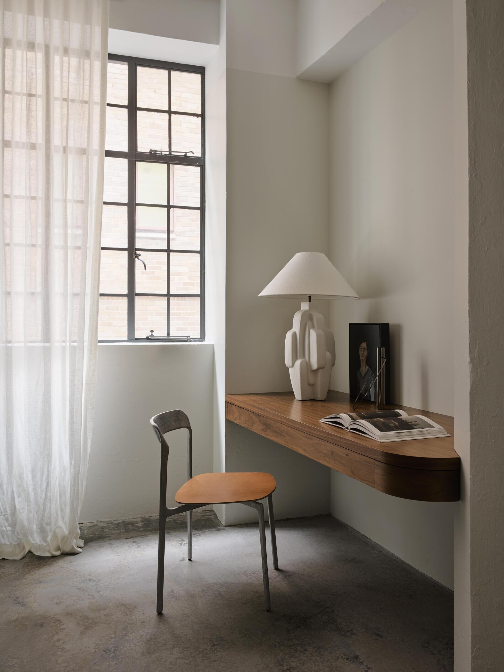 A minimalist home office nook with a floating wood desk, rounded corner, sculptural chair, and ceramic table lamp. The desk sits beside a tall black steel-framed industrial window with full-length white curtains.