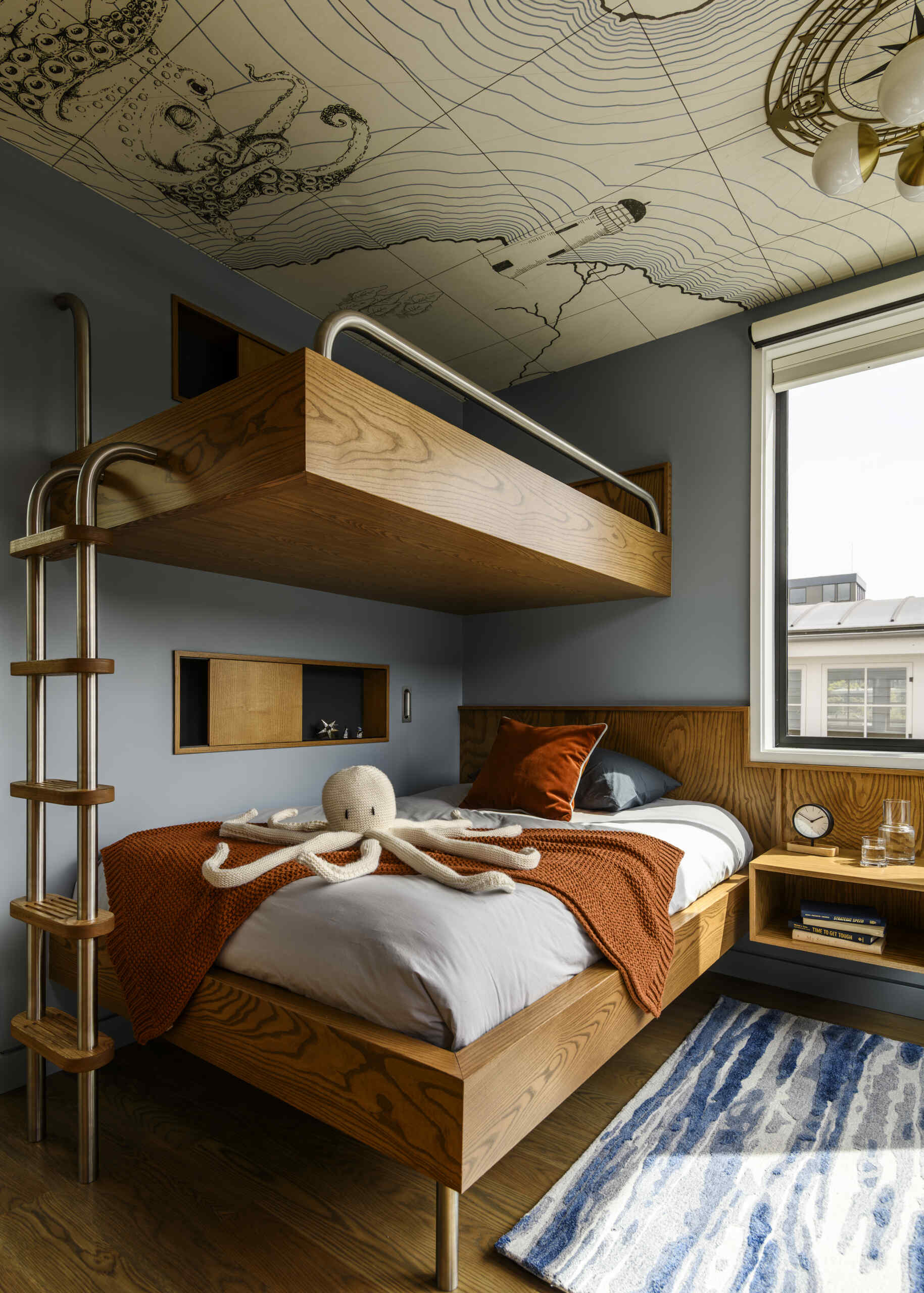 Featuring custom wood bunk beds, stainless steel railings, nautical wallpaper on the ceiling, a built-in desk, and warm orange accents, this compact bedroom combines playful design with practical storage.
