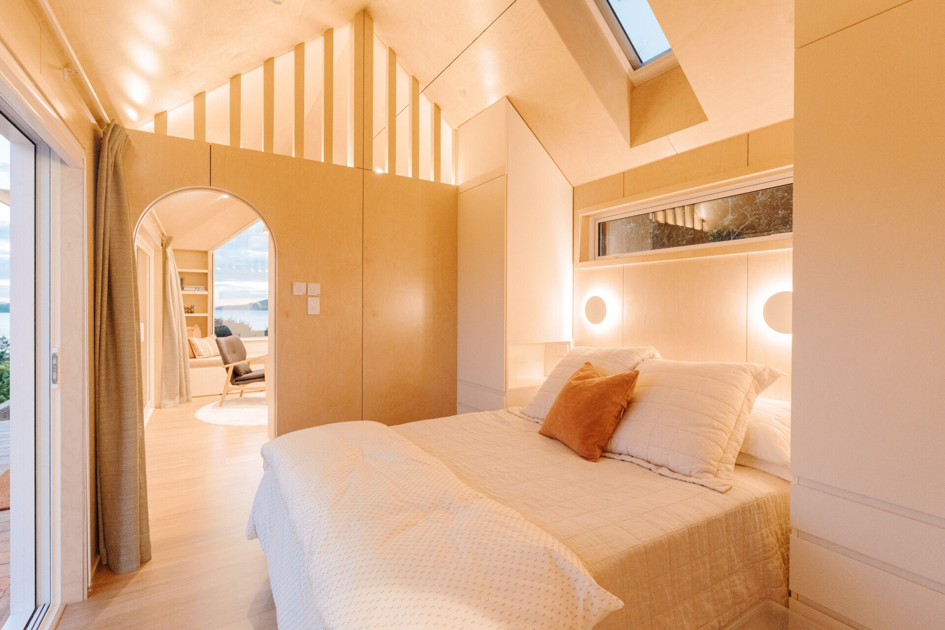 A cosy modern cabin bedroom with birch plywood walls, a skylight above the bed, custom built-in storage, warm ambient lighting and an arched doorway. Beautiful small bedroom inspiration from New Zealand.