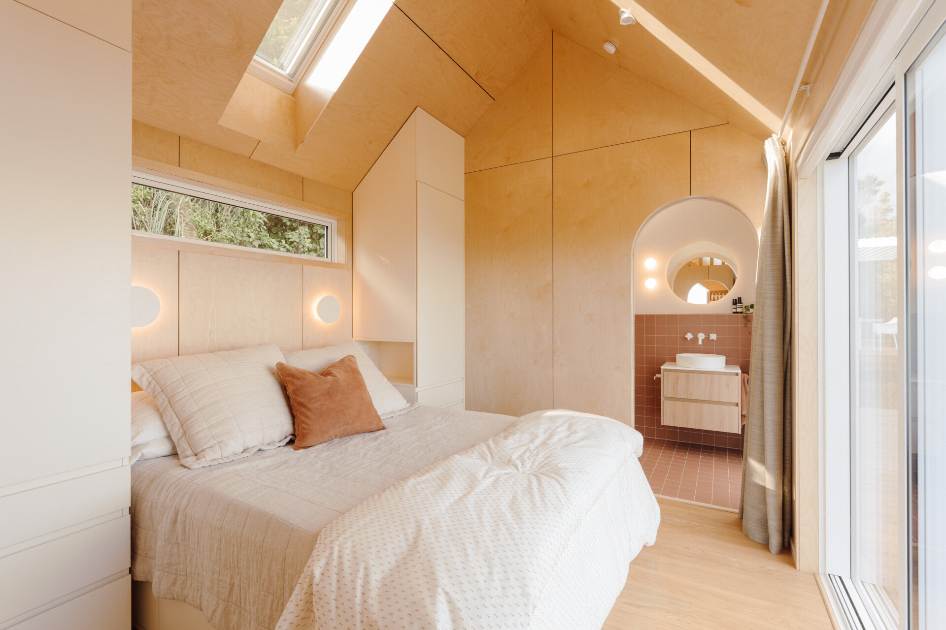 A cosy modern cabin bedroom with birch plywood walls, a skylight above the bed, custom built-in storage, warm ambient lighting and an arched doorway. Beautiful small bedroom inspiration from New Zealand.