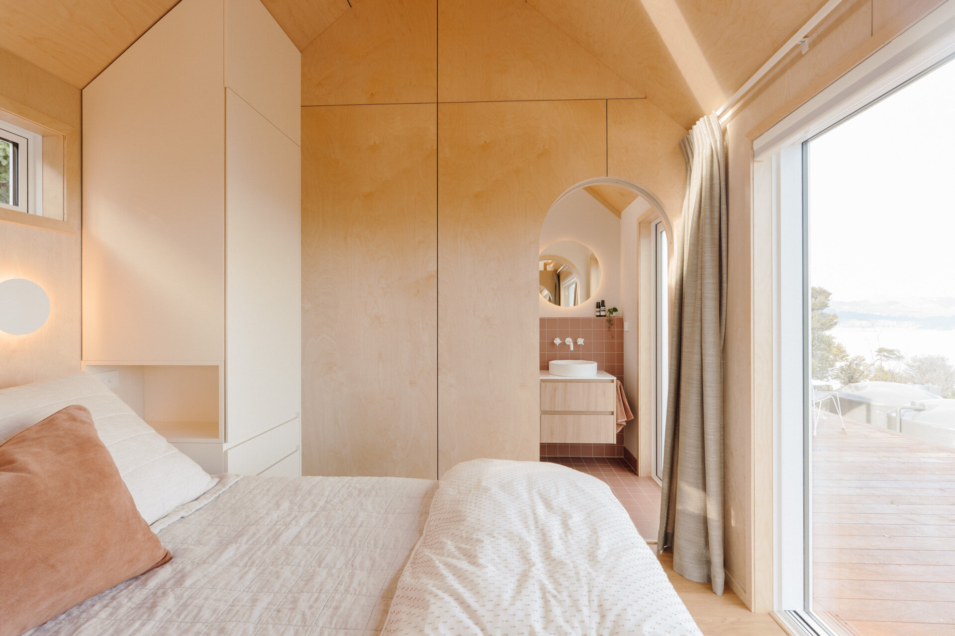 A cosy modern cabin bedroom with birch plywood walls, a skylight above the bed, custom built-in storage, warm ambient lighting and an arched doorway. Beautiful small bedroom inspiration from New Zealand.