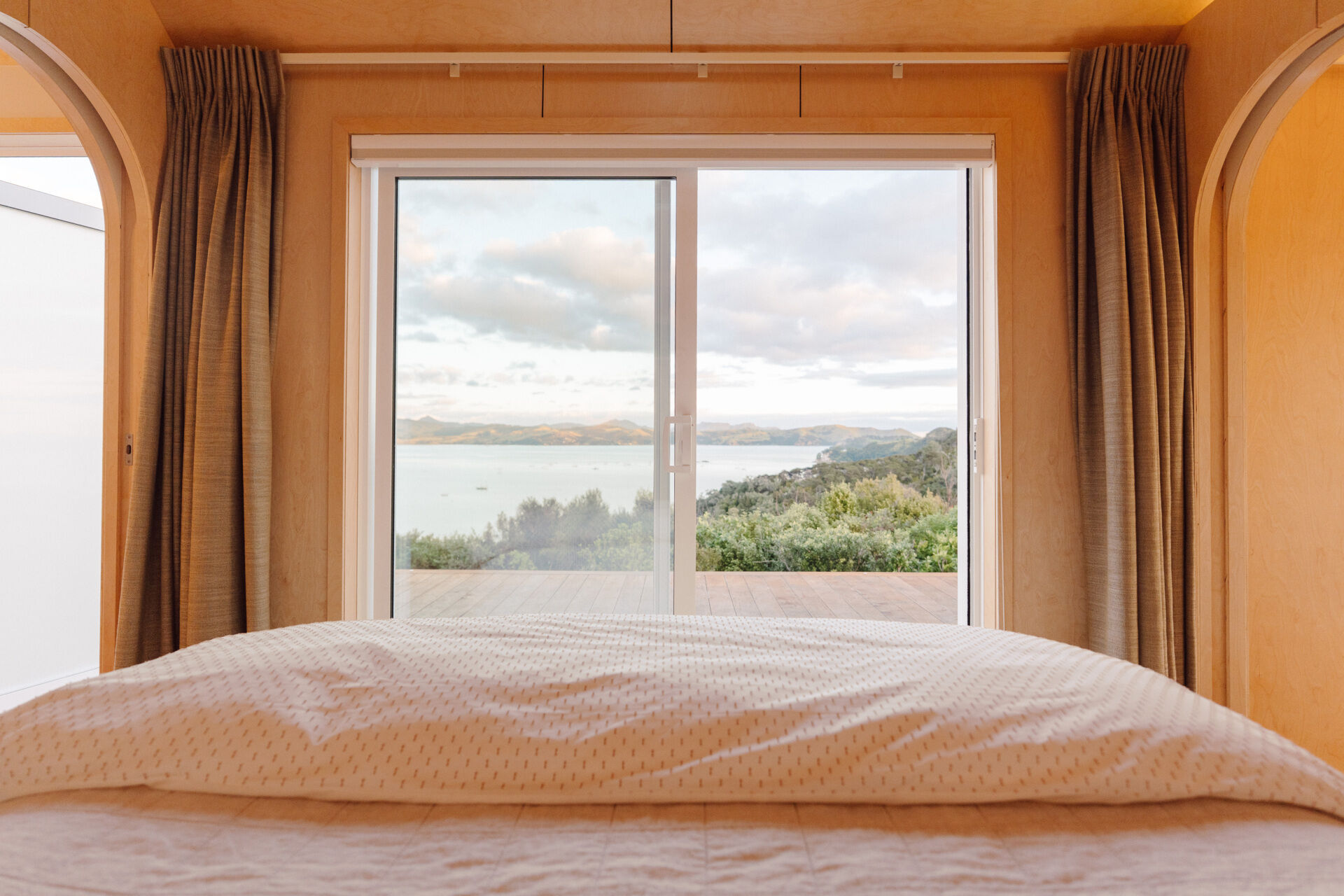 A cosy modern cabin bedroom with birch plywood walls, a skylight above the bed, custom built-in storage, warm ambient lighting and an arched doorway. Beautiful small bedroom inspiration from New Zealand.