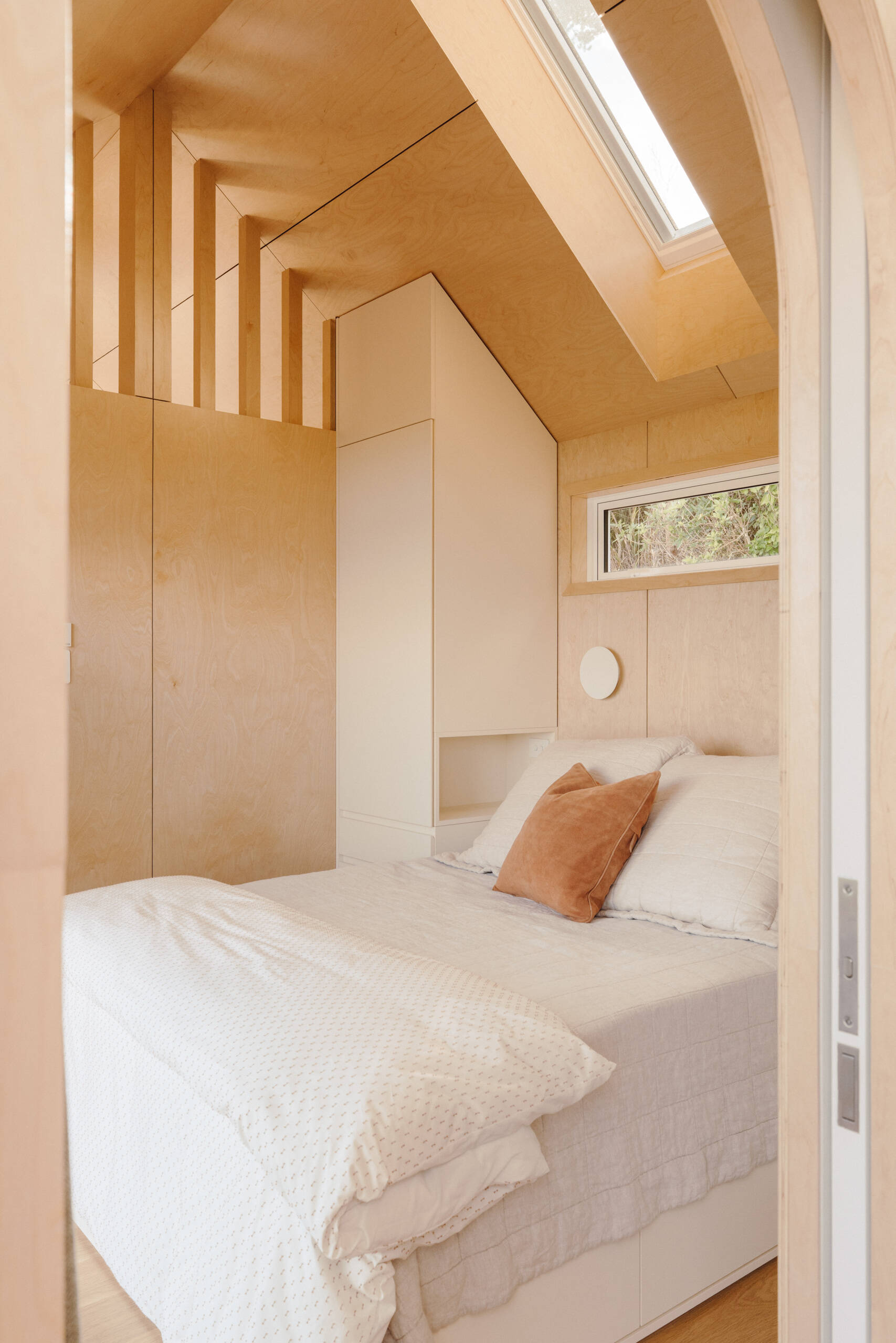 A cosy modern cabin bedroom with birch plywood walls, a skylight above the bed, custom built-in storage, warm ambient lighting and an arched doorway. Beautiful small bedroom inspiration from New Zealand.