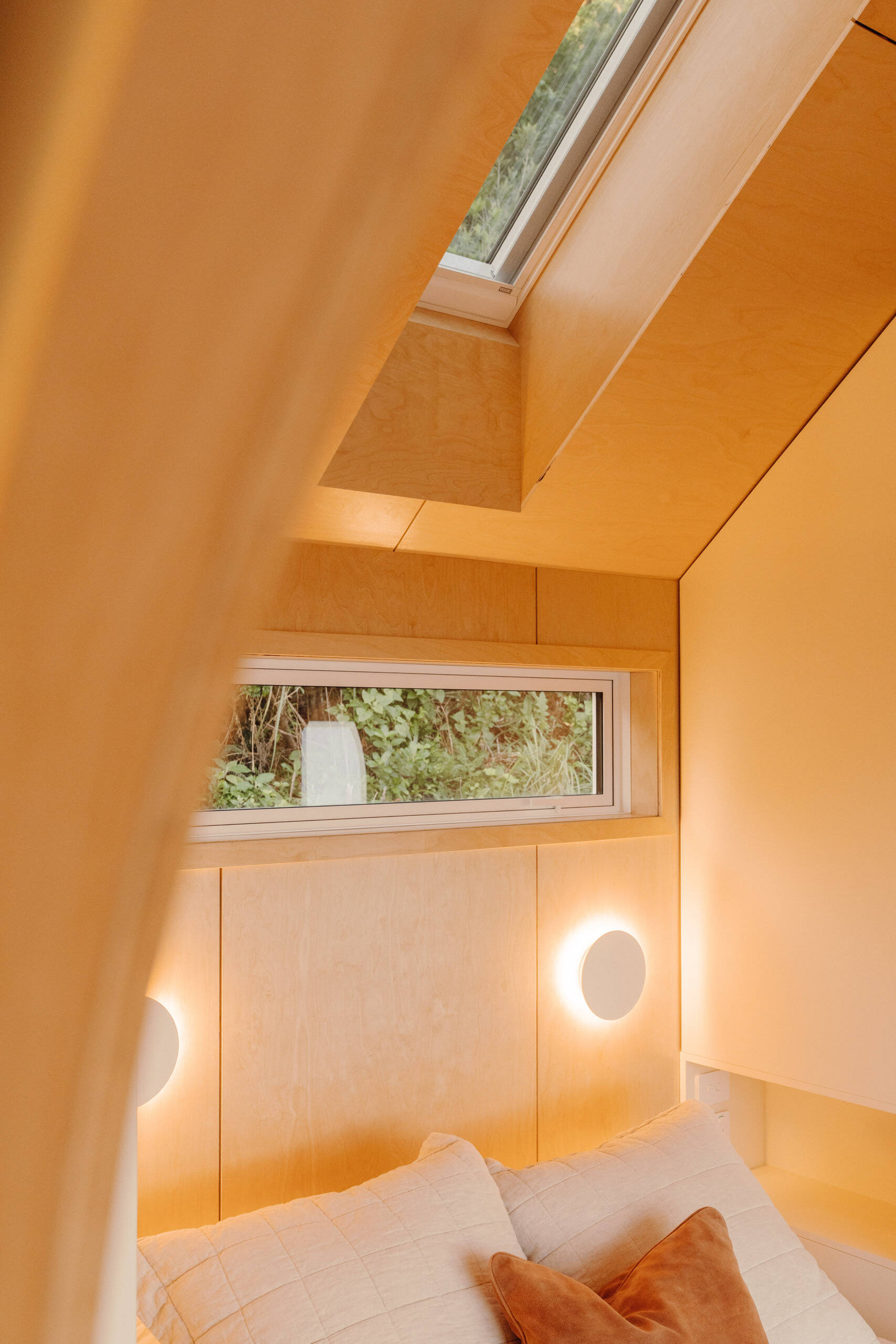 A cosy modern cabin bedroom with birch plywood walls, a skylight above the bed, custom built-in storage, warm ambient lighting and an arched doorway. Beautiful small bedroom inspiration from New Zealand.