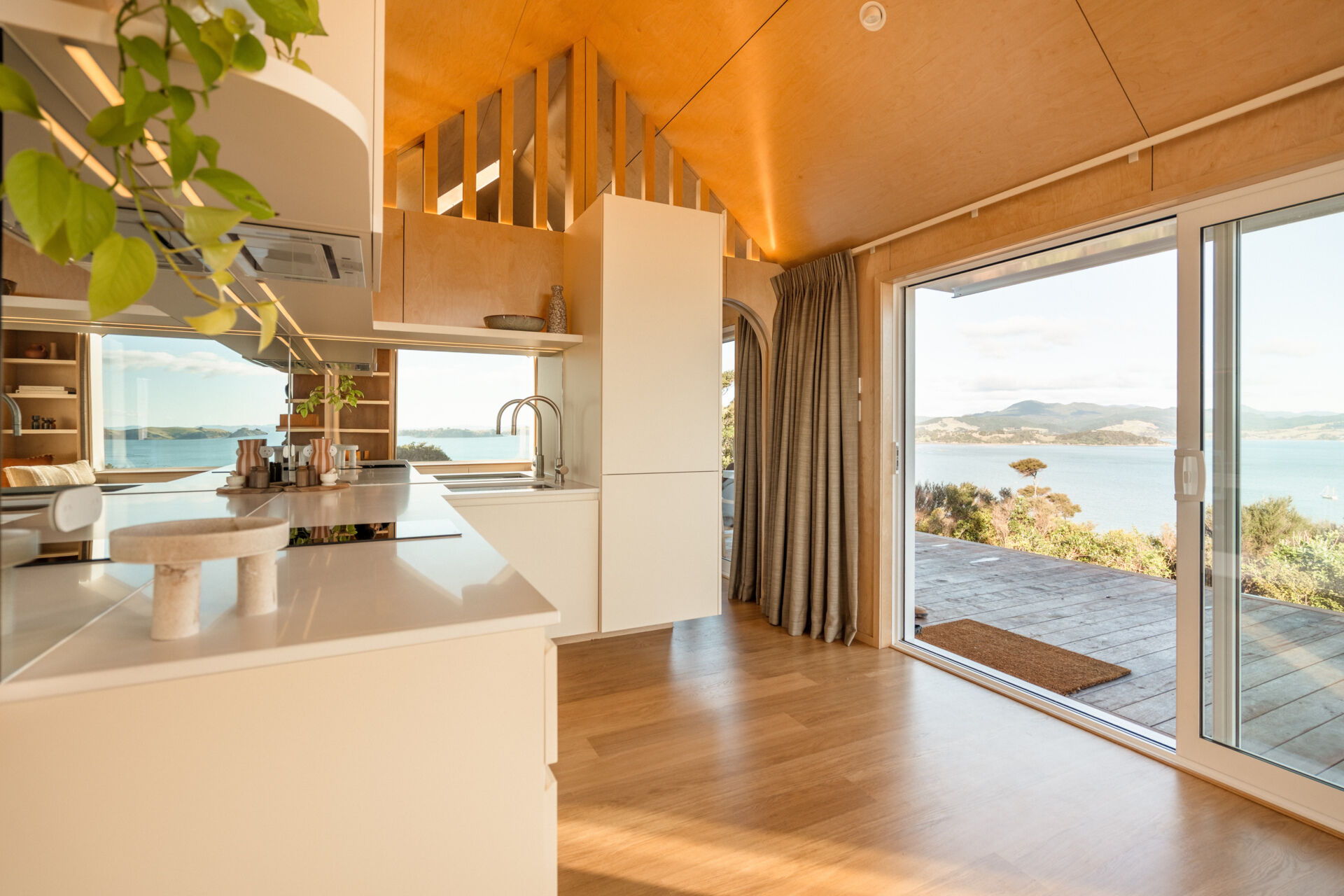 Explore a compact modern kitchen with white custom cabinets, mirrored backsplash, integrated appliances and warm birch plywood walls. Clever kitchenette inspiration from Arc's 30m² New Zealand cabin.
