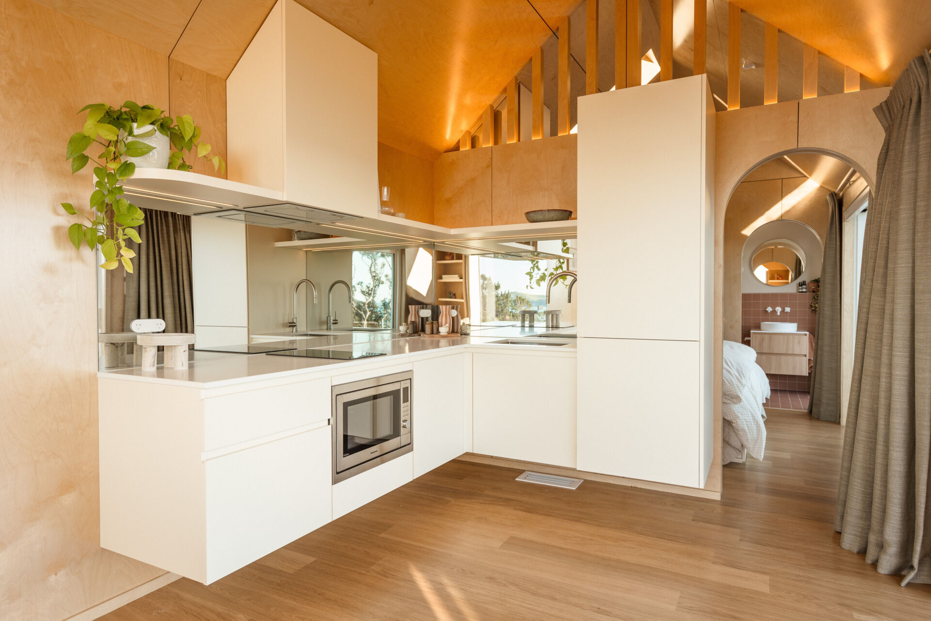 Explore a compact modern kitchen with white custom cabinets, mirrored backsplash, integrated appliances and warm birch plywood walls. Clever kitchenette inspiration from Arc's 30m² New Zealand cabin.