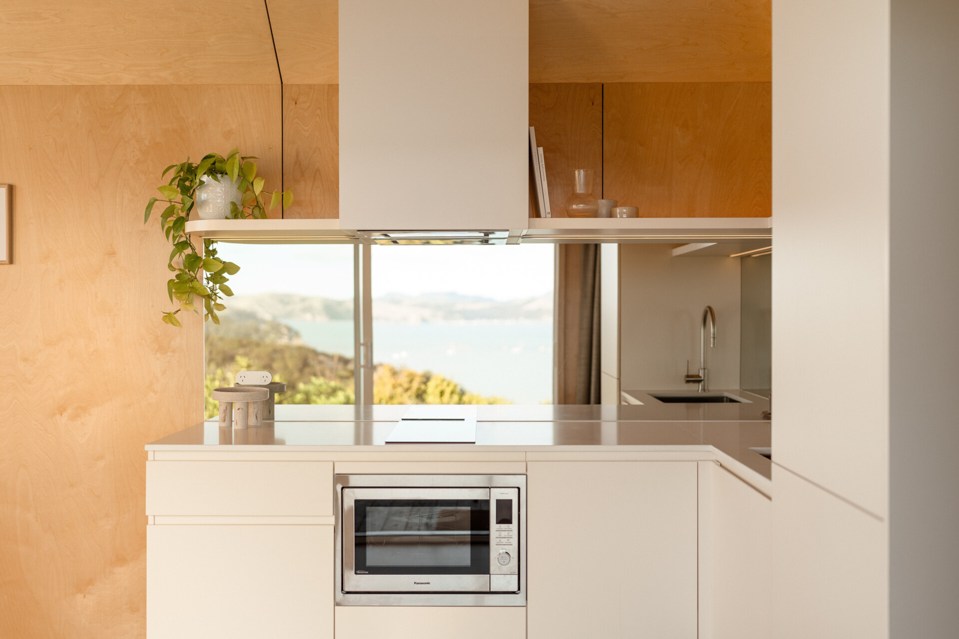 Explore a compact modern kitchen with white custom cabinets, mirrored backsplash, integrated appliances and warm birch plywood walls. Clever kitchenette inspiration from Arc's 30m² New Zealand cabin.