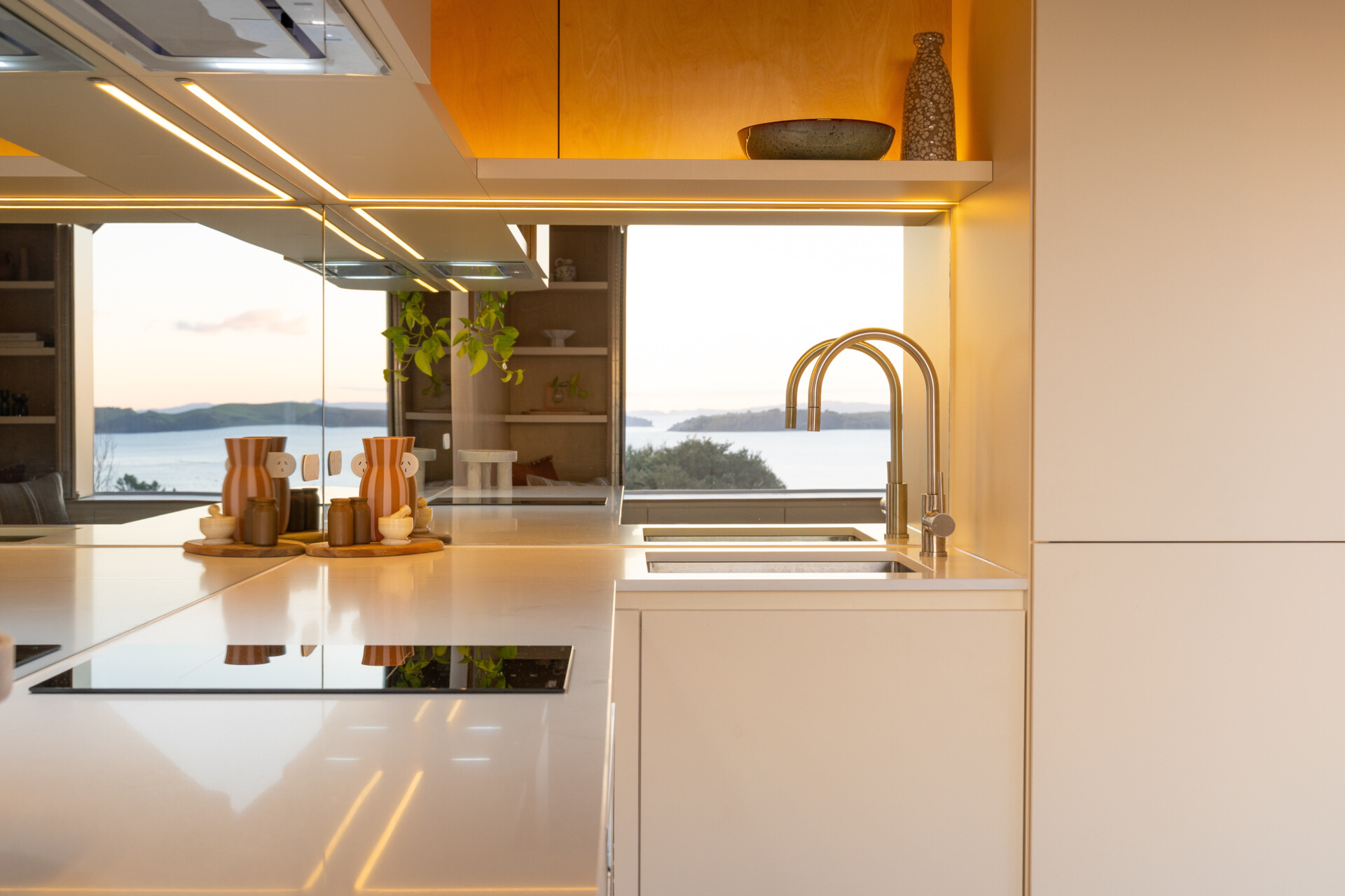 Explore a compact modern kitchen with white custom cabinets, mirrored backsplash, integrated appliances and warm birch plywood walls. Clever kitchenette inspiration from Arc's 30m² New Zealand cabin.