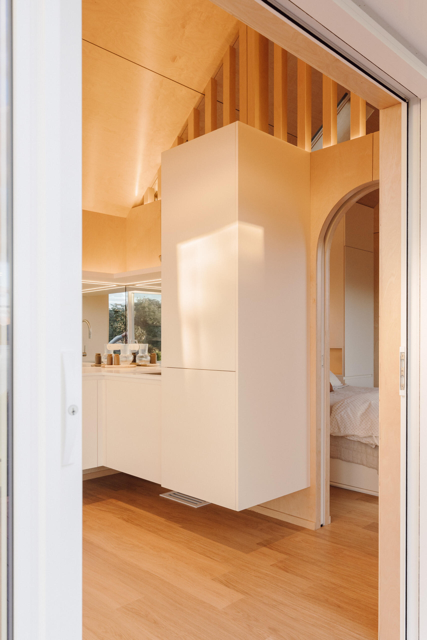 Explore a compact modern kitchen with white custom cabinets, mirrored backsplash, integrated appliances and warm birch plywood walls. Clever kitchenette inspiration from Arc's 30m² New Zealand cabin.