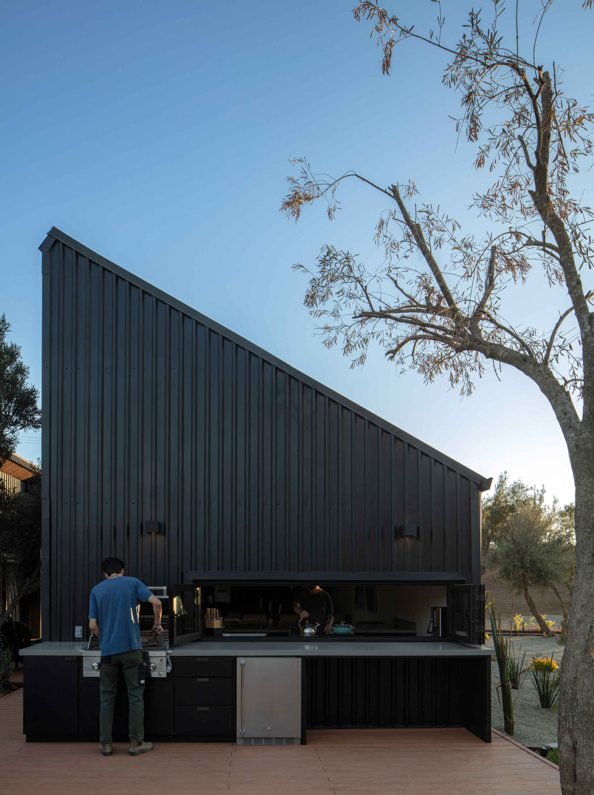 Discover the outdoor kitchen at Serentha Cabin by ORTO STUDIO in Baja California, Mexico. A built-in stainless steel barbecue, black cabinets, spacious countertops, and a pass-through kitchen window make this modern cabin ideal for outdoor cooking and entertaining.