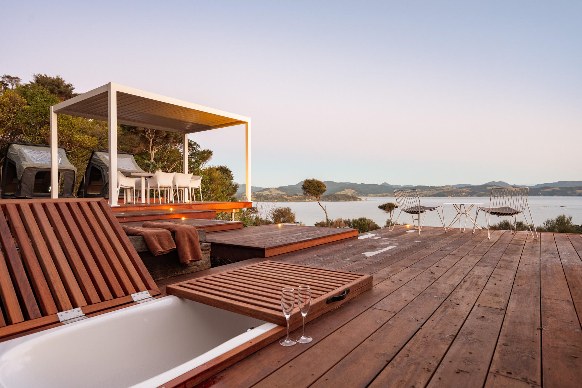 Explore the outdoor living spaces of a modern Coromandel cabin, featuring expansive timber decking, a sunken outdoor bathtub, covered dining pavilion, glamping tents and spectacular coastal views. Inspiration for small holiday homes and outdoor entertaining.