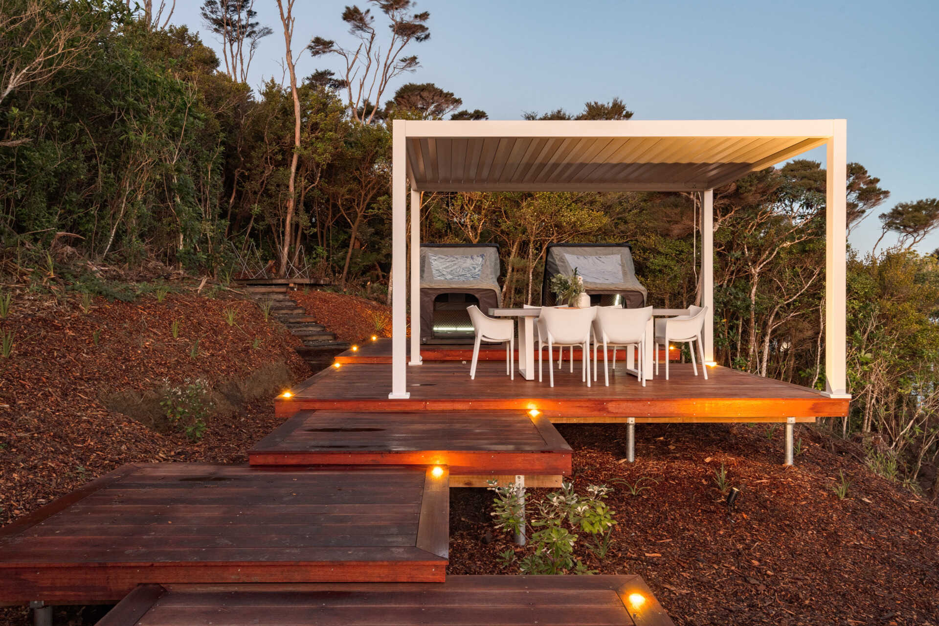 Explore the outdoor living spaces of a modern Coromandel cabin, featuring expansive timber decking, a sunken outdoor bathtub, covered dining pavilion, glamping tents and spectacular coastal views. Inspiration for small holiday homes and outdoor entertaining.