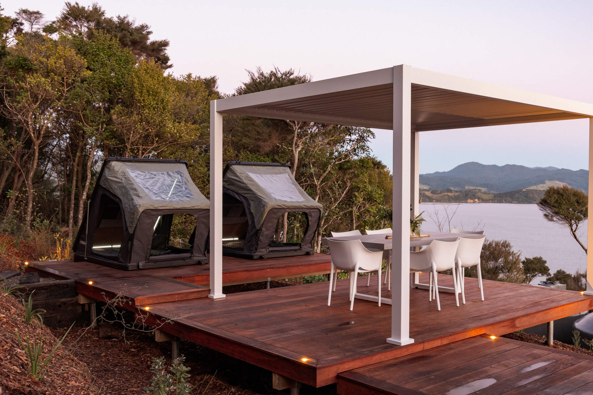 Explore the outdoor living spaces of a modern Coromandel cabin, featuring expansive timber decking, a sunken outdoor bathtub, covered dining pavilion, glamping tents and spectacular coastal views. Inspiration for small holiday homes and outdoor entertaining.