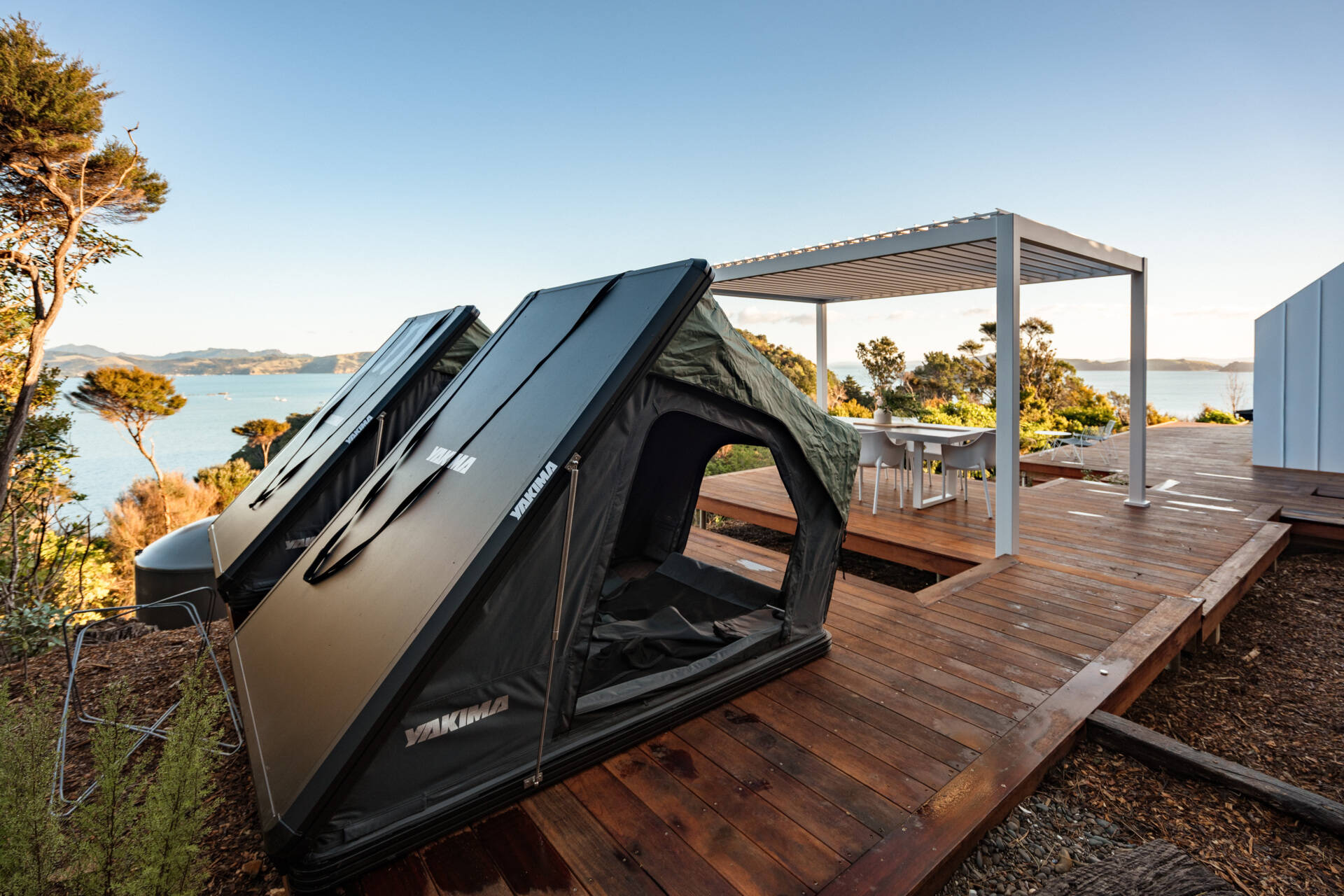 Explore the outdoor living spaces of a modern Coromandel cabin, featuring expansive timber decking, a sunken outdoor bathtub, covered dining pavilion, glamping tents and spectacular coastal views. Inspiration for small holiday homes and outdoor entertaining.