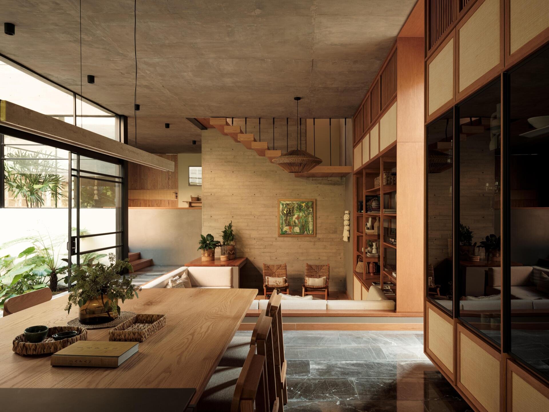 Explore a modern dining room and kitchen in Casa Akina, Mexico, featuring a solid wood dining table, custom timber cabinetry, a dark stone kitchen island, minimalist lighting, and large windows overlooking tropical greenery.