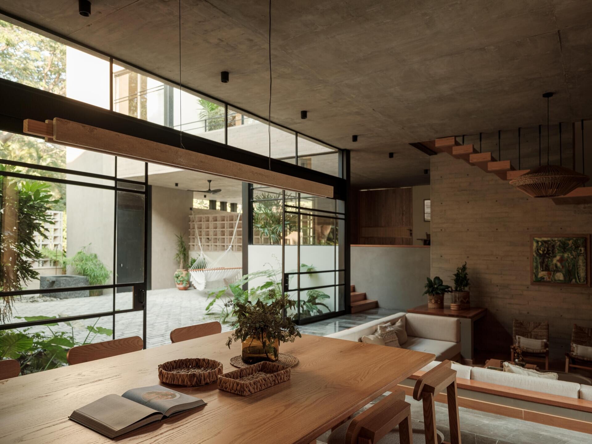 Explore a modern dining room and kitchen in Casa Akina, Mexico, featuring a solid wood dining table, custom timber cabinetry, a dark stone kitchen island, minimalist lighting, and large windows overlooking tropical greenery.