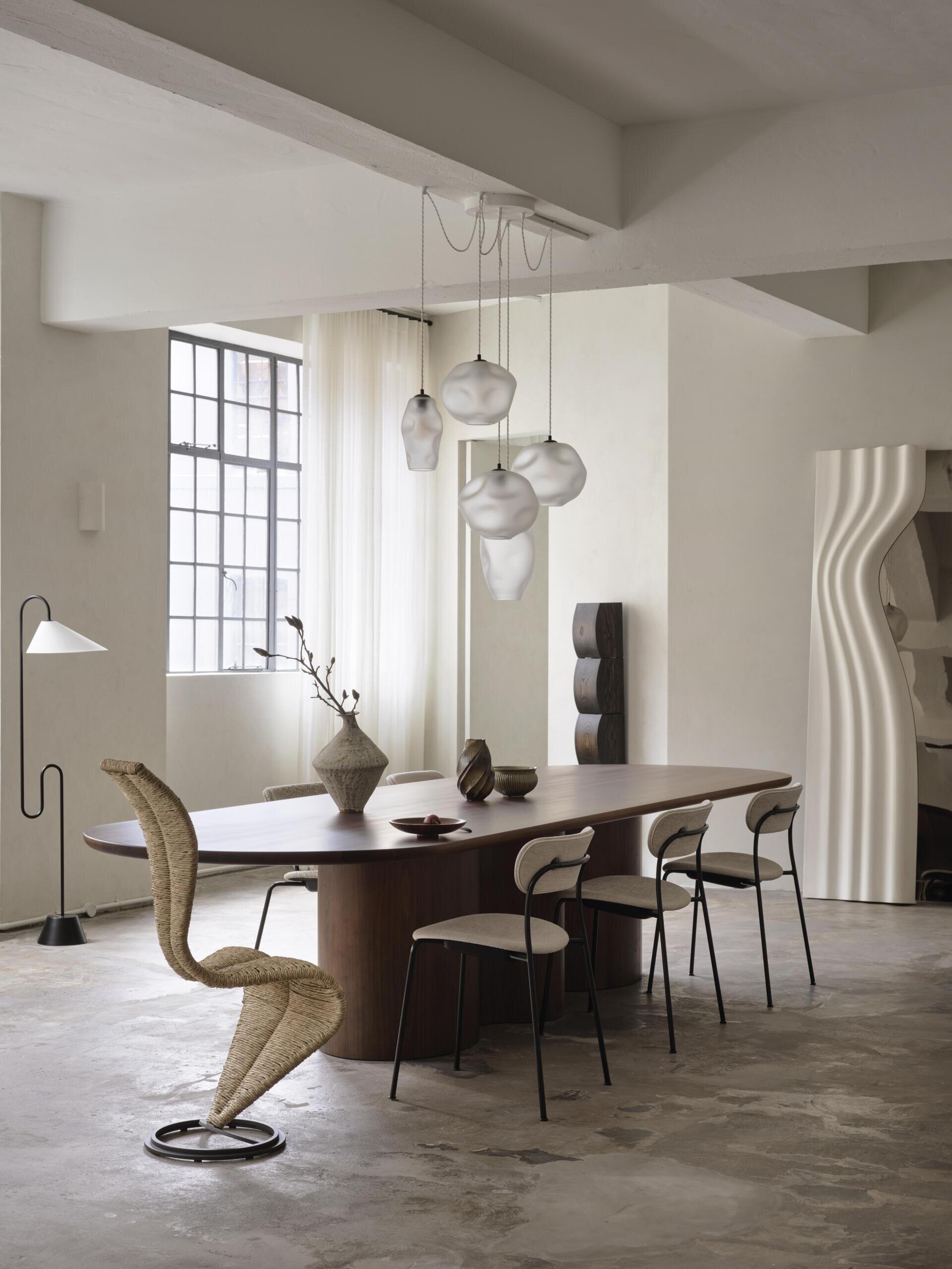 A contemporary dining room with a long dark wood table, woven sculptural chair, upholstered dining chairs, and a cluster of organic-shaped white pendant lights. Black steel-framed industrial windows and a curved white doorway add architectural character.