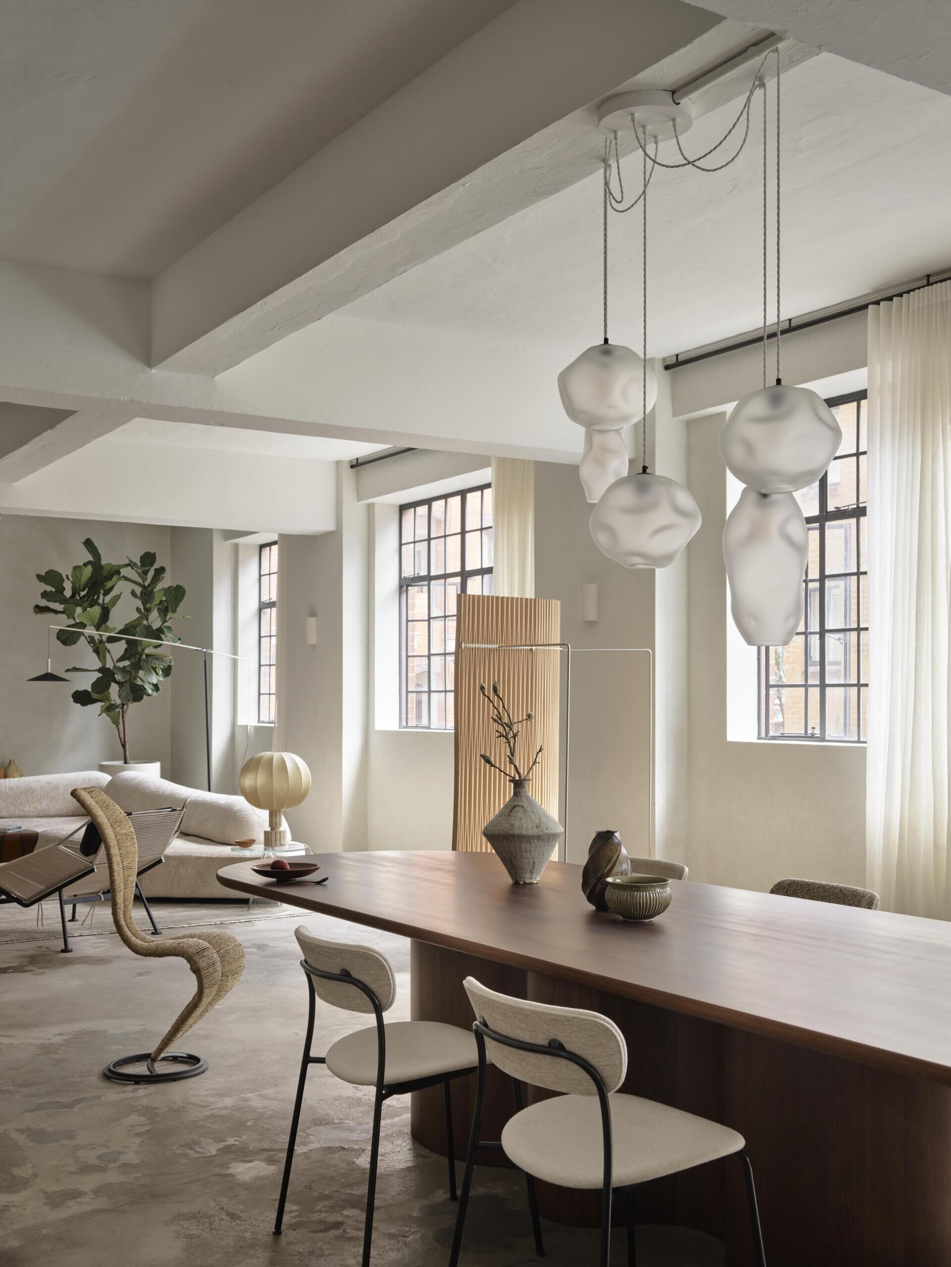 An open-plan dining and living space with a large wood dining table, sculptural white glass pendant lights, and original industrial-style windows. Soft white curtains and a neutral sofa introduce warmth to the apartment's concrete interior.
