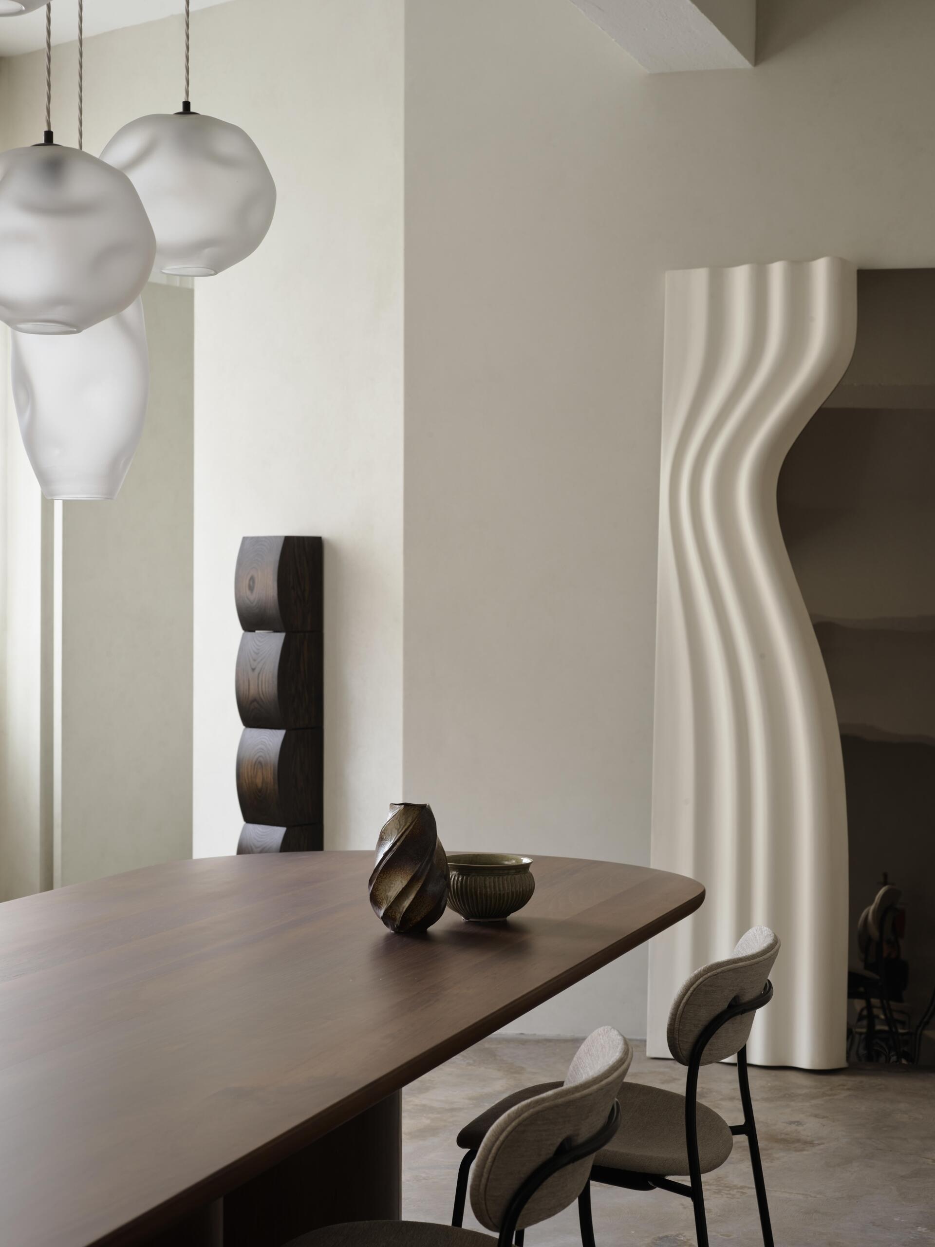 A minimalist dining area featuring a dark wood table, ceramic vessels, soft white pendant lights, and an unusual sculptural doorway with flowing curved edges. A tall dark wood sculpture adds contrast to the pale interior.