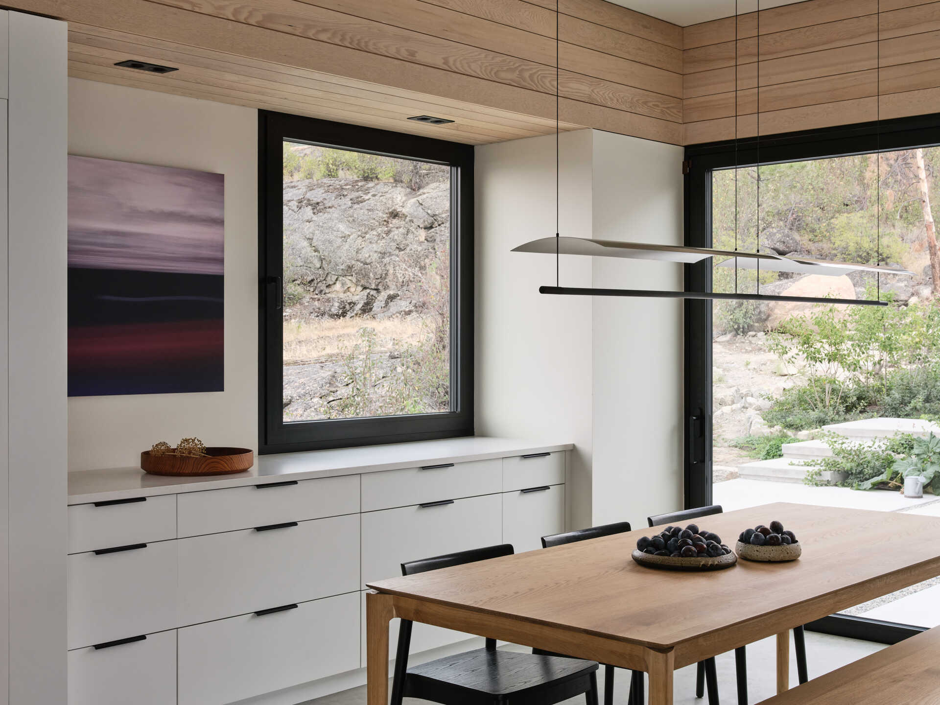 A modern dining room with a natural wood table, black dining chairs, white built-in storage, and sculptural pendant lighting. Large black-framed windows and a glass door frame views of the rocky landscape outside.